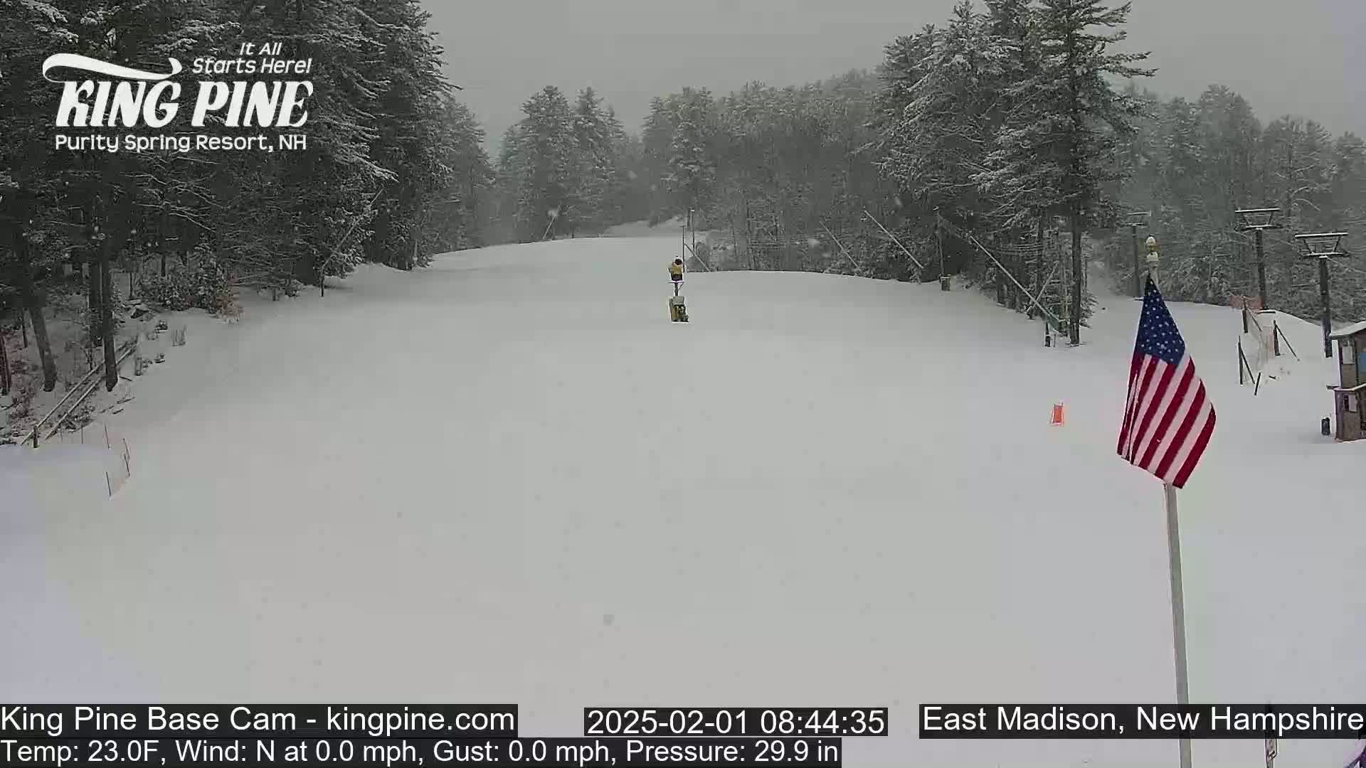 A snow-covered ski slope is seen with a person skiing down it, under a heavy snowfall, with a small American flag in the lower right.