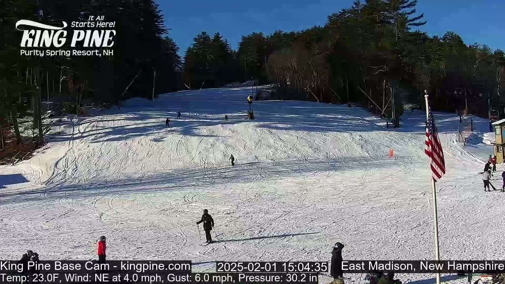 A snow-covered ski slope with several skiers and snowboarders, under a clear sunny sky.