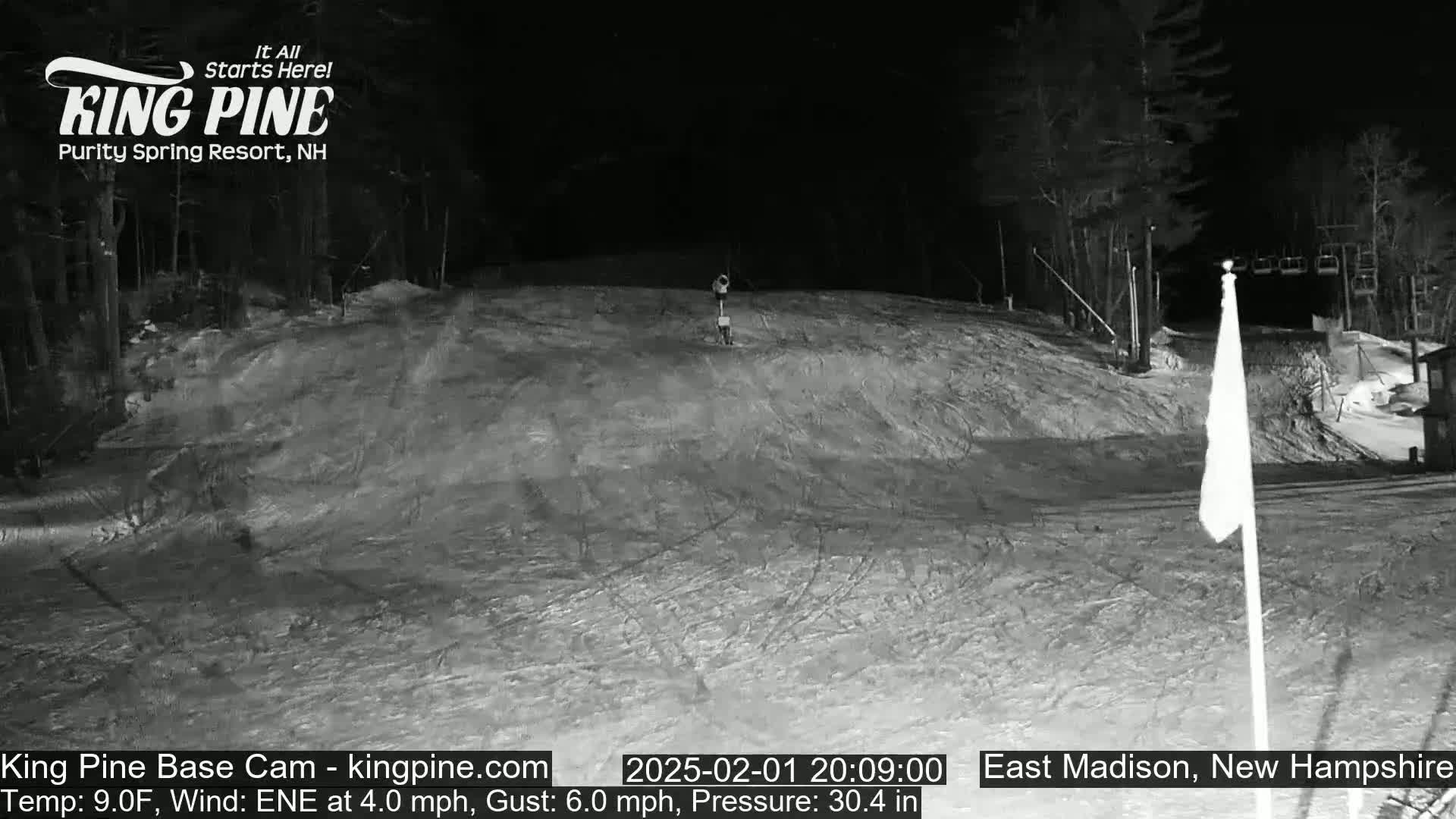 A snow-covered ski slope at night, illuminated by artificial light, with a ski lift visible in the background and a lone figure in the distance.