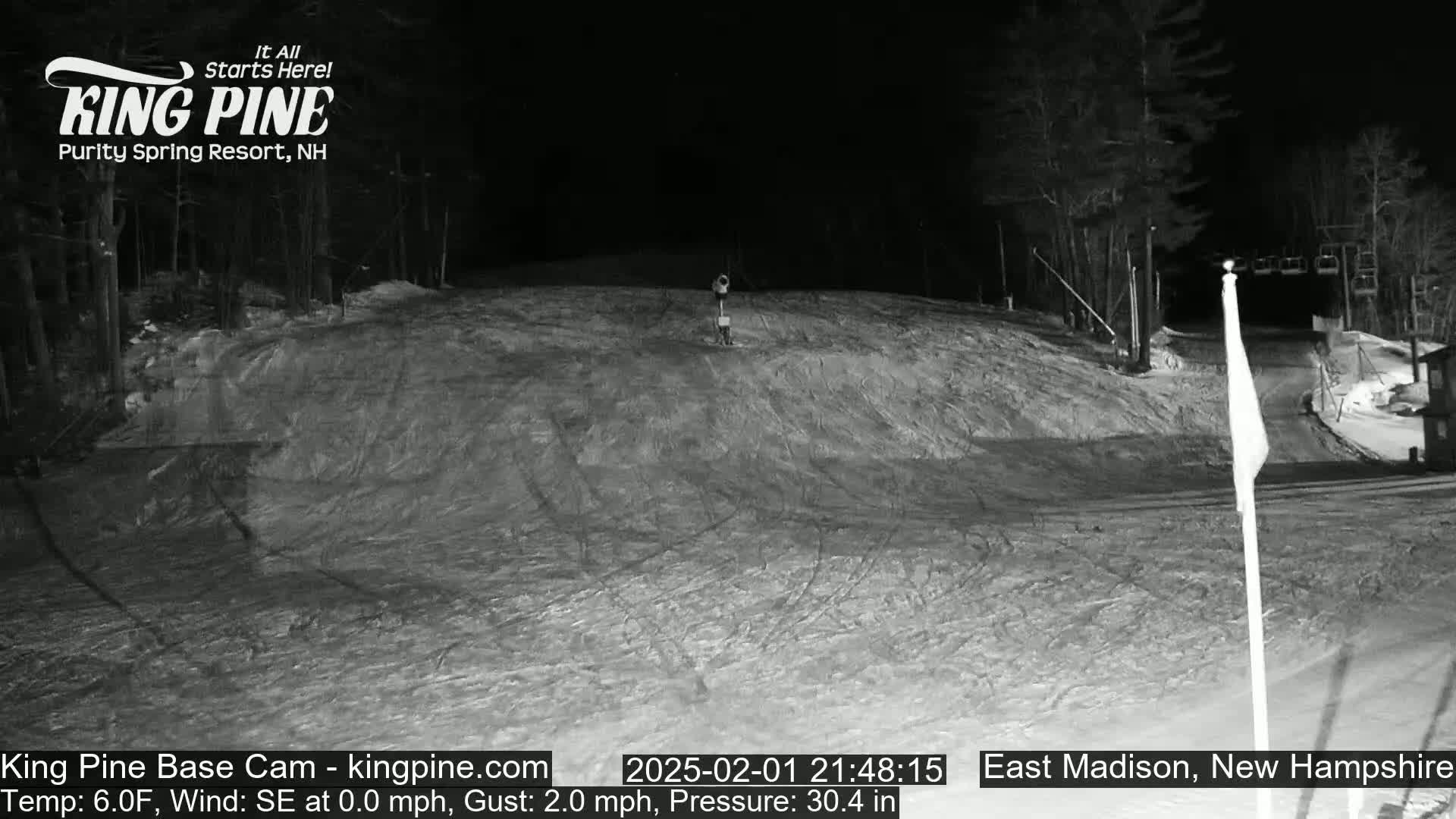 A nighttime, snow-covered ski slope with a chairlift visible in the background under a dark sky.