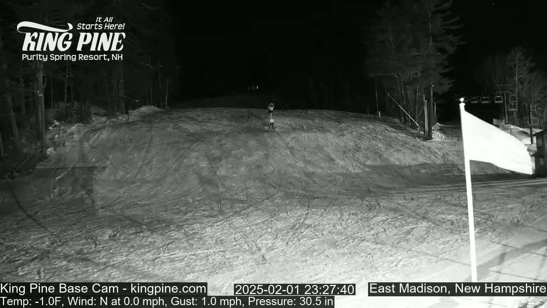 A snow-covered ski slope at night, illuminated by artificial light, with a single figure visible in the distance.