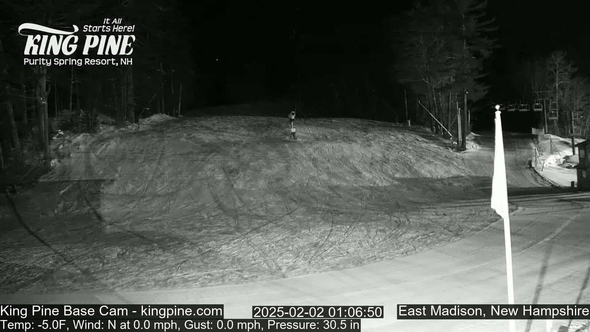 A snow-covered ski slope at night, illuminated by artificial light, with a chairlift visible in the background.