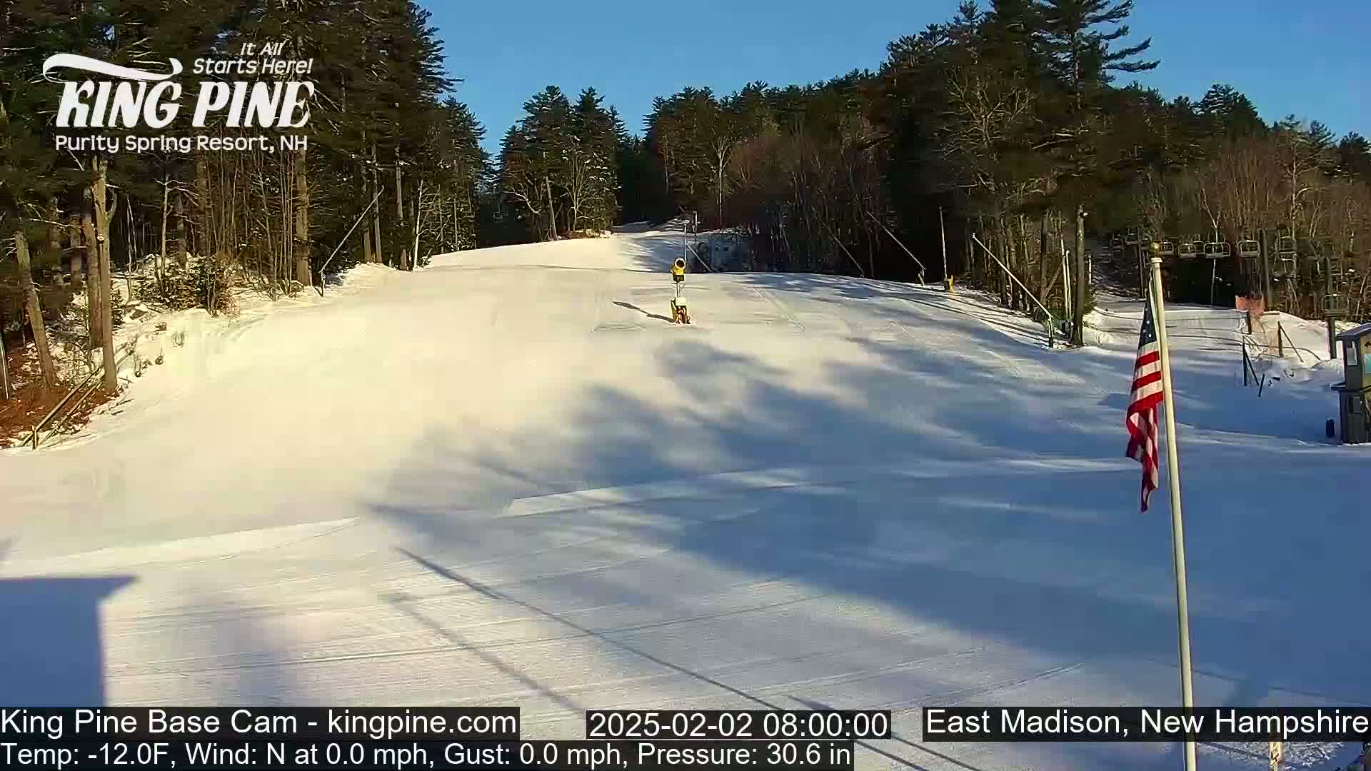 A snow-covered ski slope is visible under a clear blue sky, with trees lining the edges and a small American flag planted near a building in the lower right.