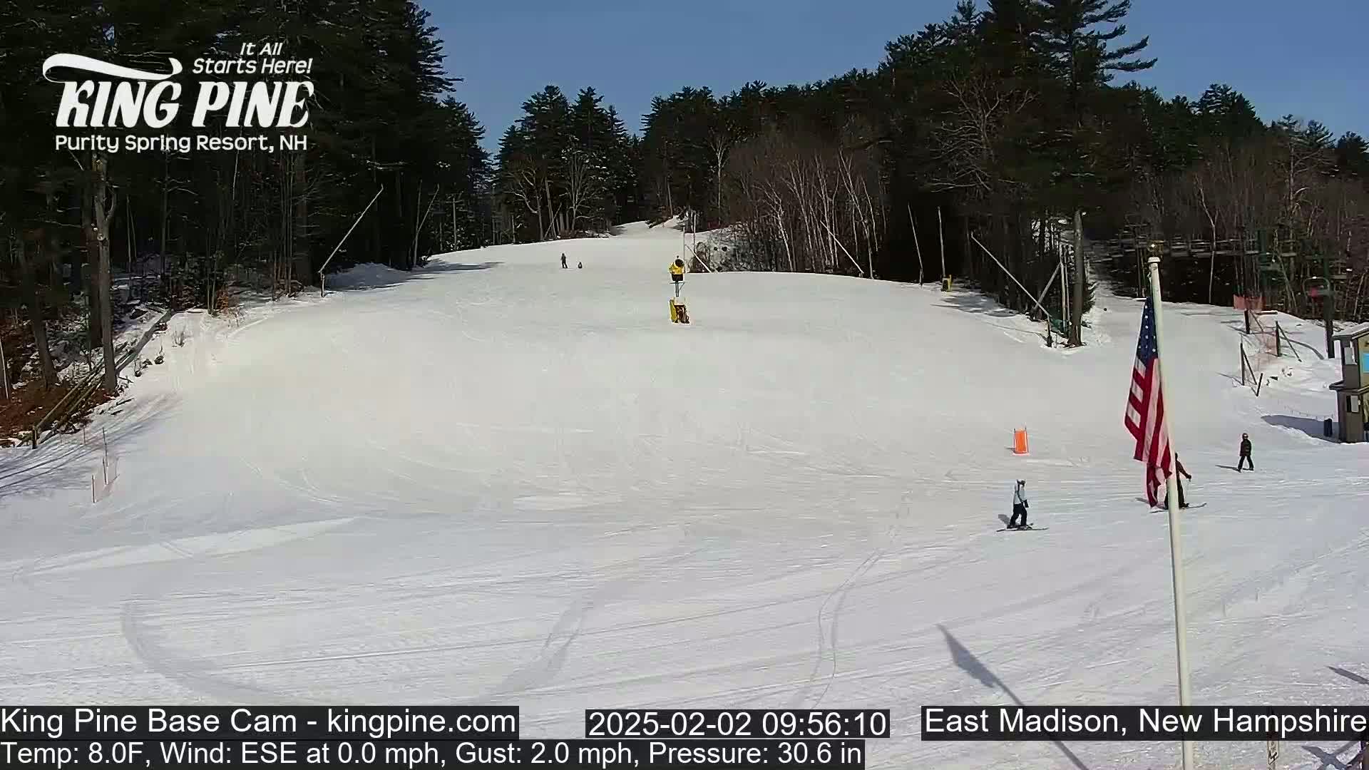 A snowy ski slope on a sunny day with several skiers and snowboarders visible, surrounded by a wooded area.