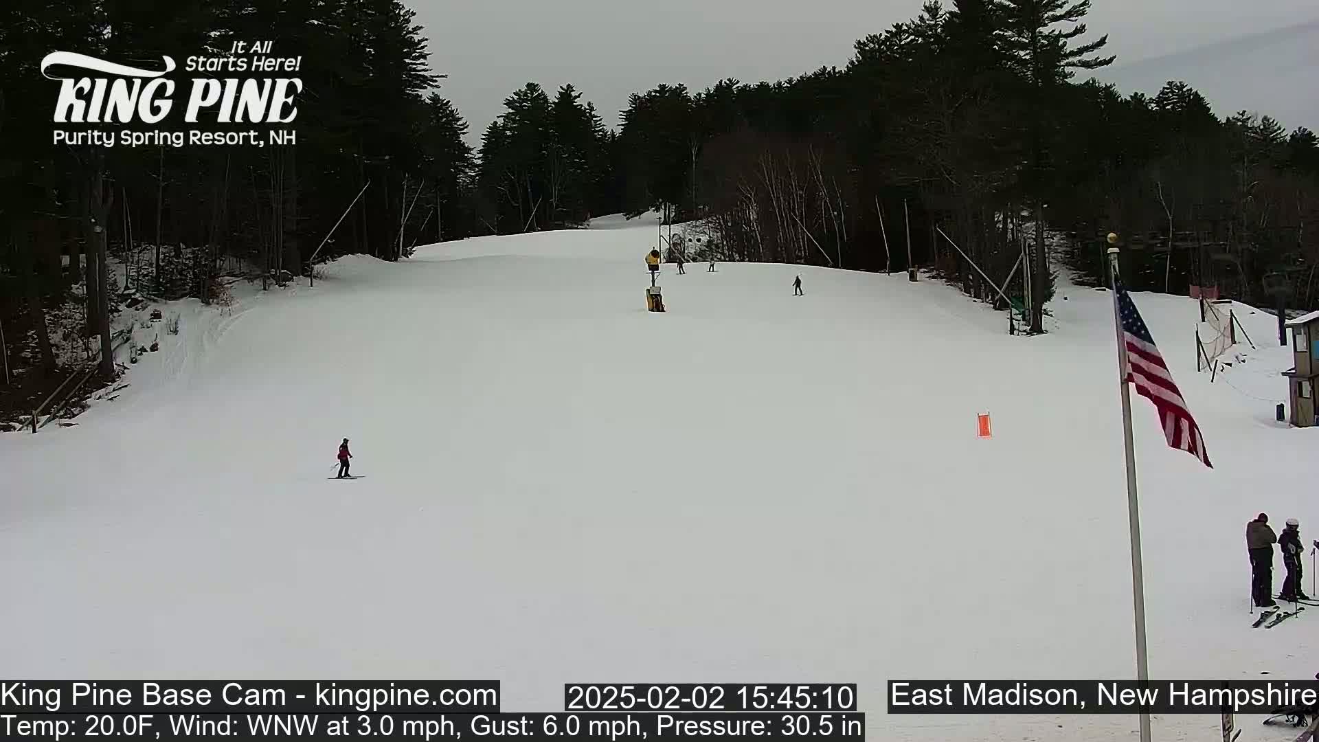 A snowy ski slope with several skiers, a flagpole with an American flag, and a wooded area in the background under an overcast sky.