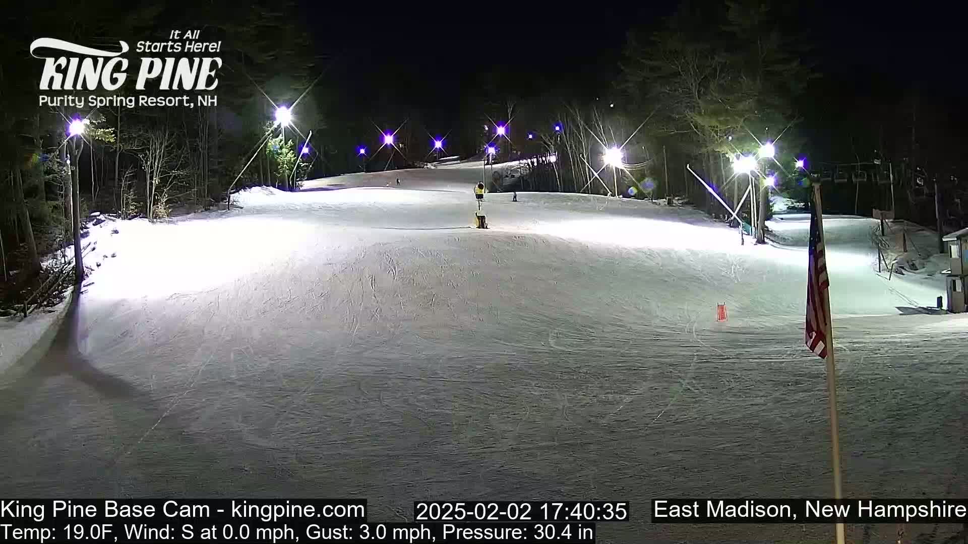 A snow-covered ski slope is brightly lit at night, with several people skiing or snowboarding down the hill.