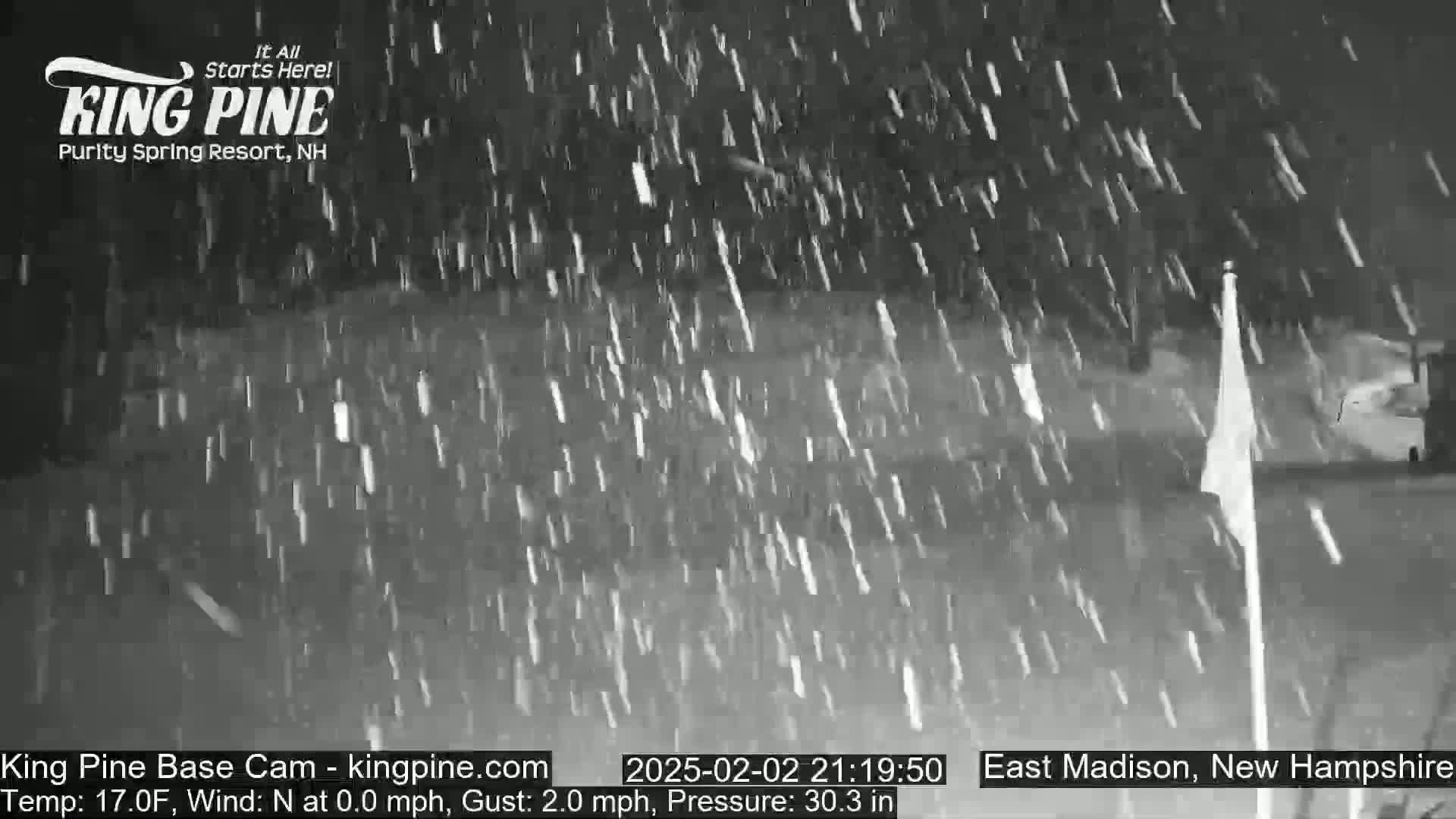 Heavy snow falls at night on a mostly dark landscape with a flagpole visible in the foreground.