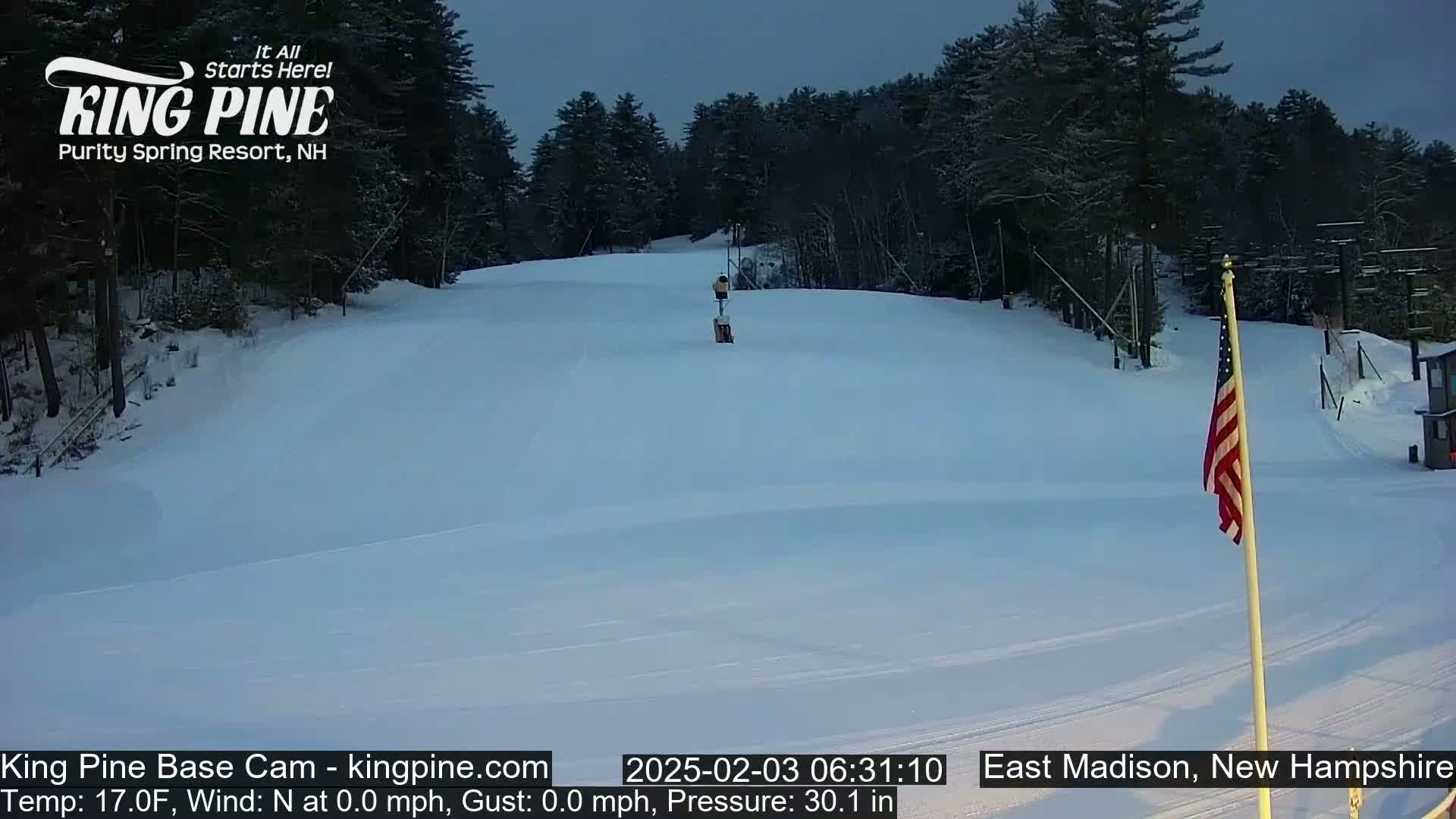 A snow-covered ski slope is visible under a twilight sky, with a small structure and an American flag in the foreground.