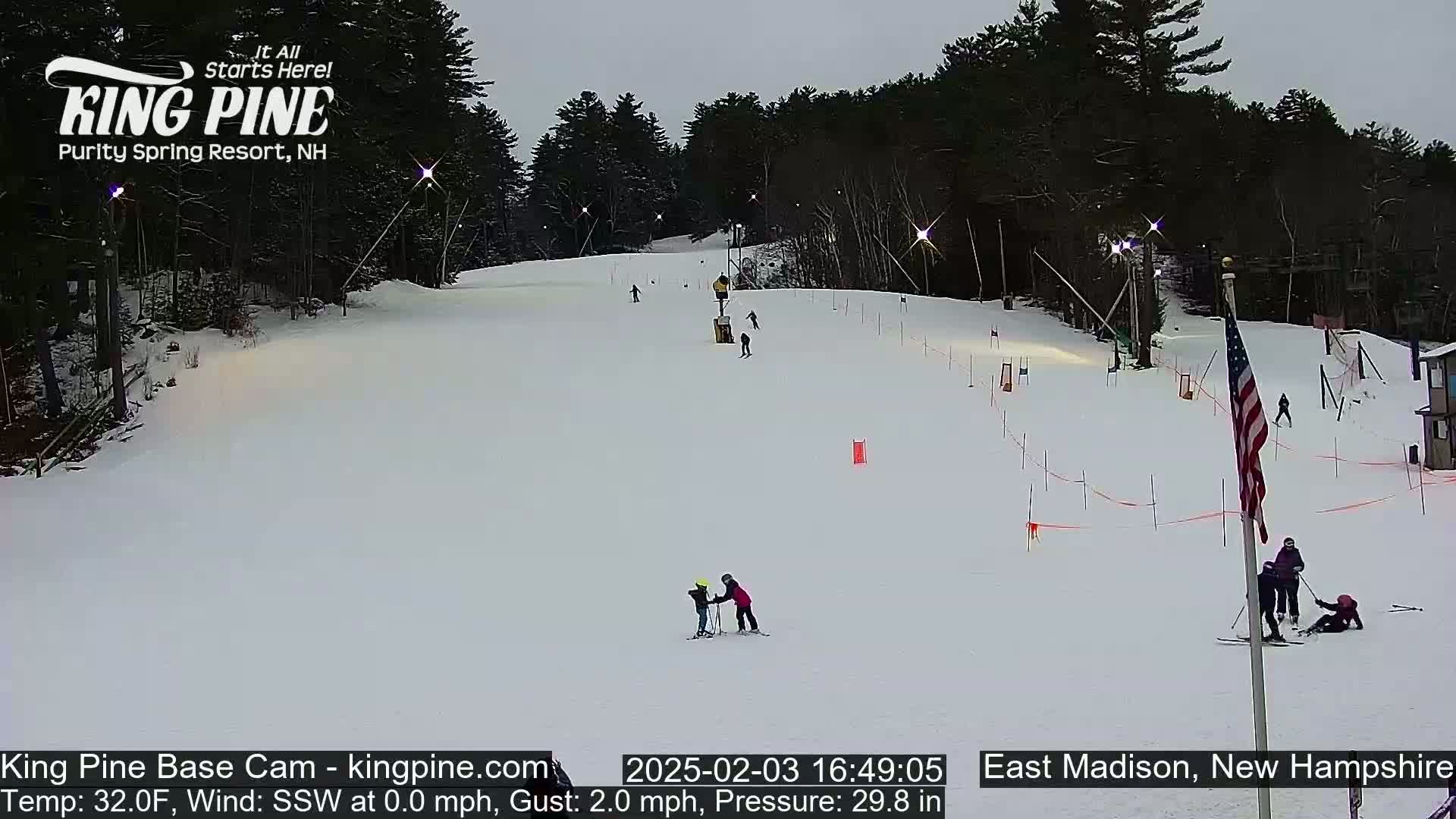 A snow-covered ski slope with several skiers and snowboarders visible under an overcast sky.