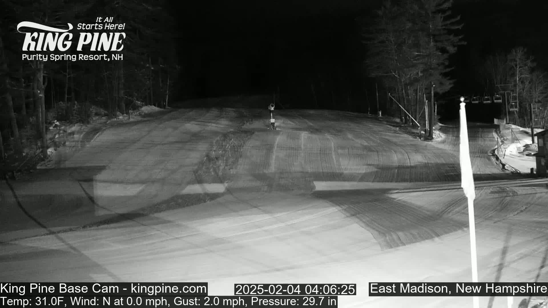 A nighttime, snow-covered ski slope is illuminated, with a calm wind and trees visible in the background.