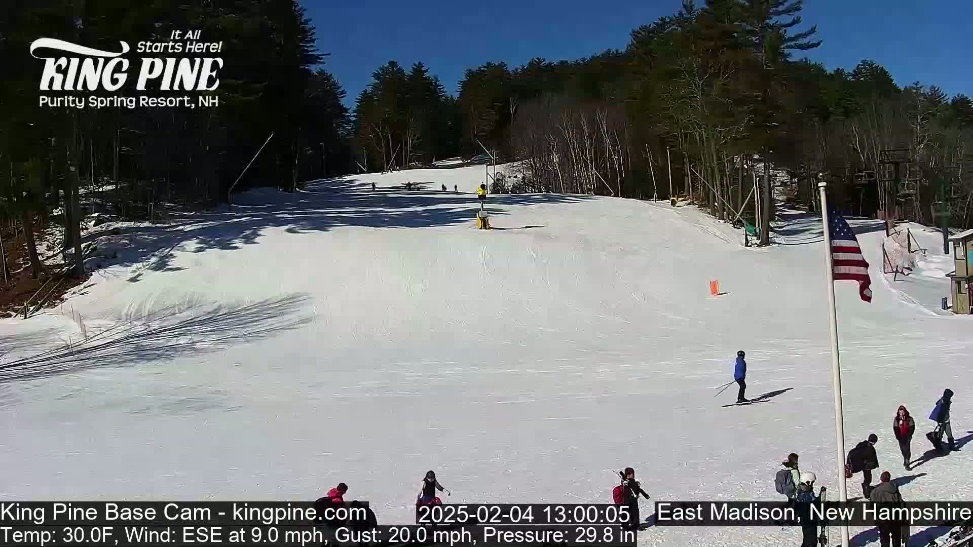 A snow-covered ski slope with several skiers and onlookers under a clear blue sky.