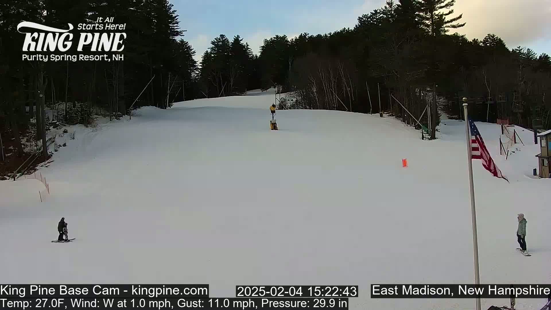 A snowy ski slope with several skiers and snowboarders, under a mostly clear sky.