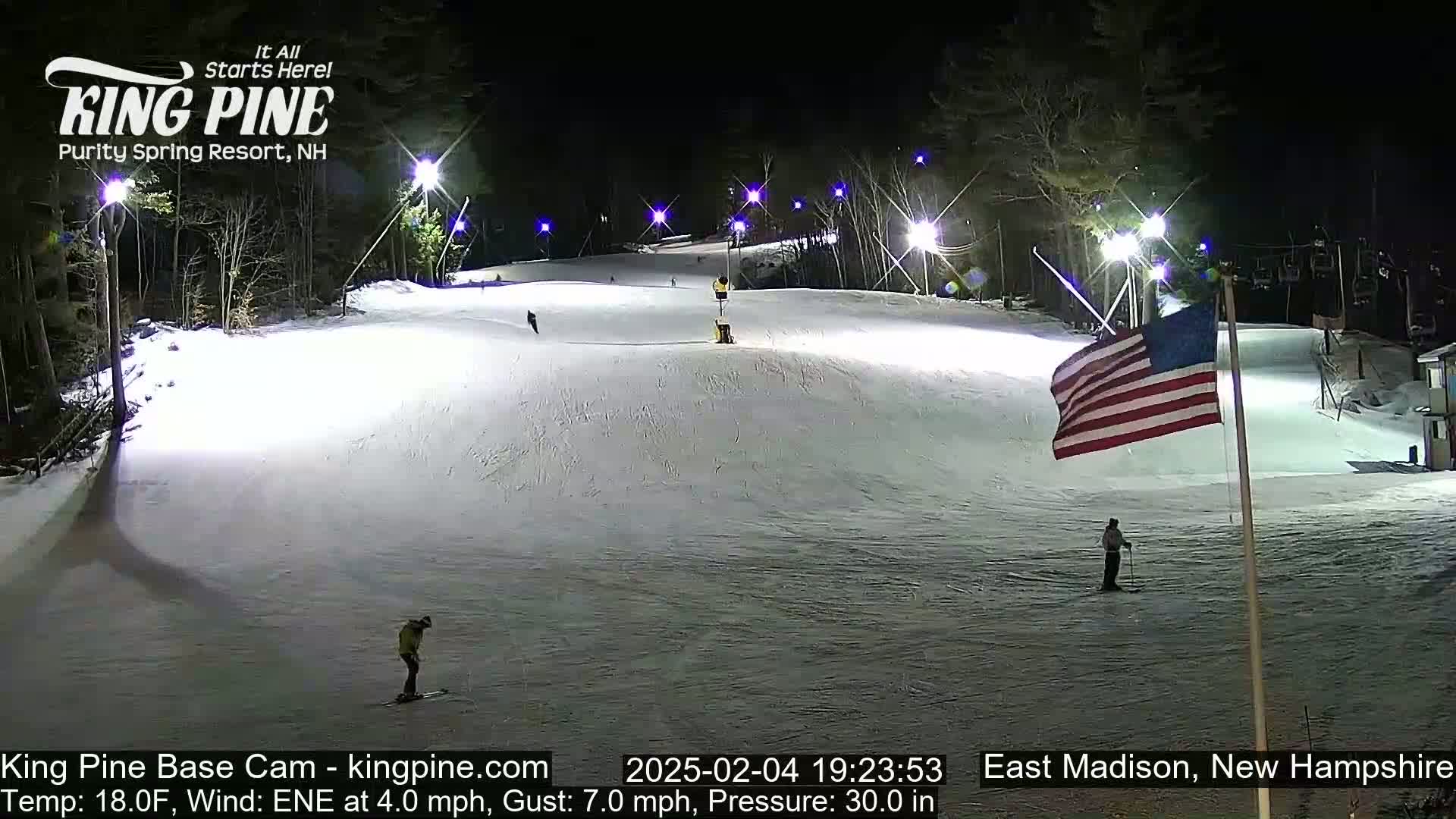 A nighttime view of a snow-covered ski slope illuminated by lights, with several skiers present and an American flag waving in the background.