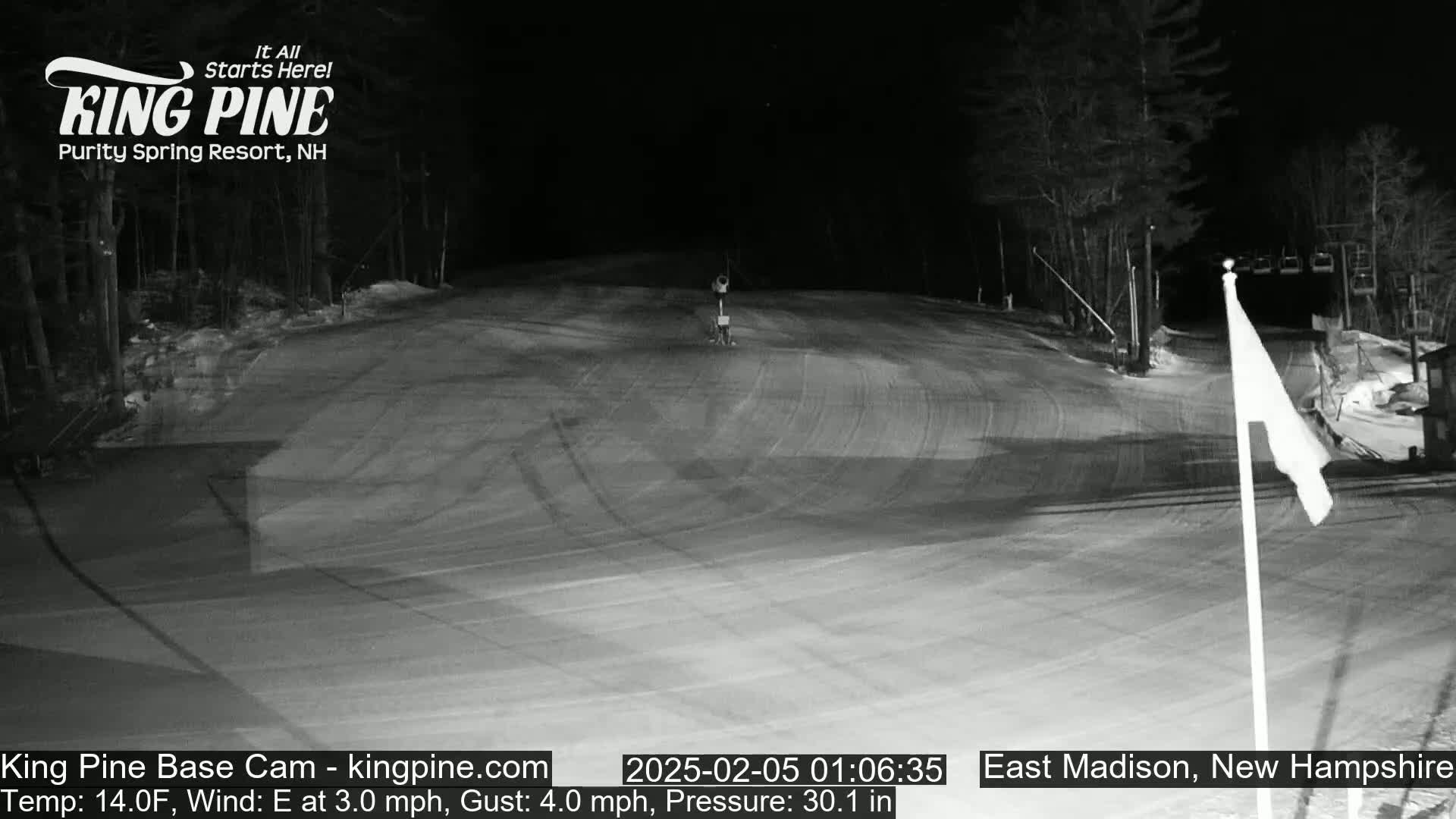 A nighttime, snow-covered ski slope is mostly empty except for a single person in the distance, with a gentle east wind blowing.