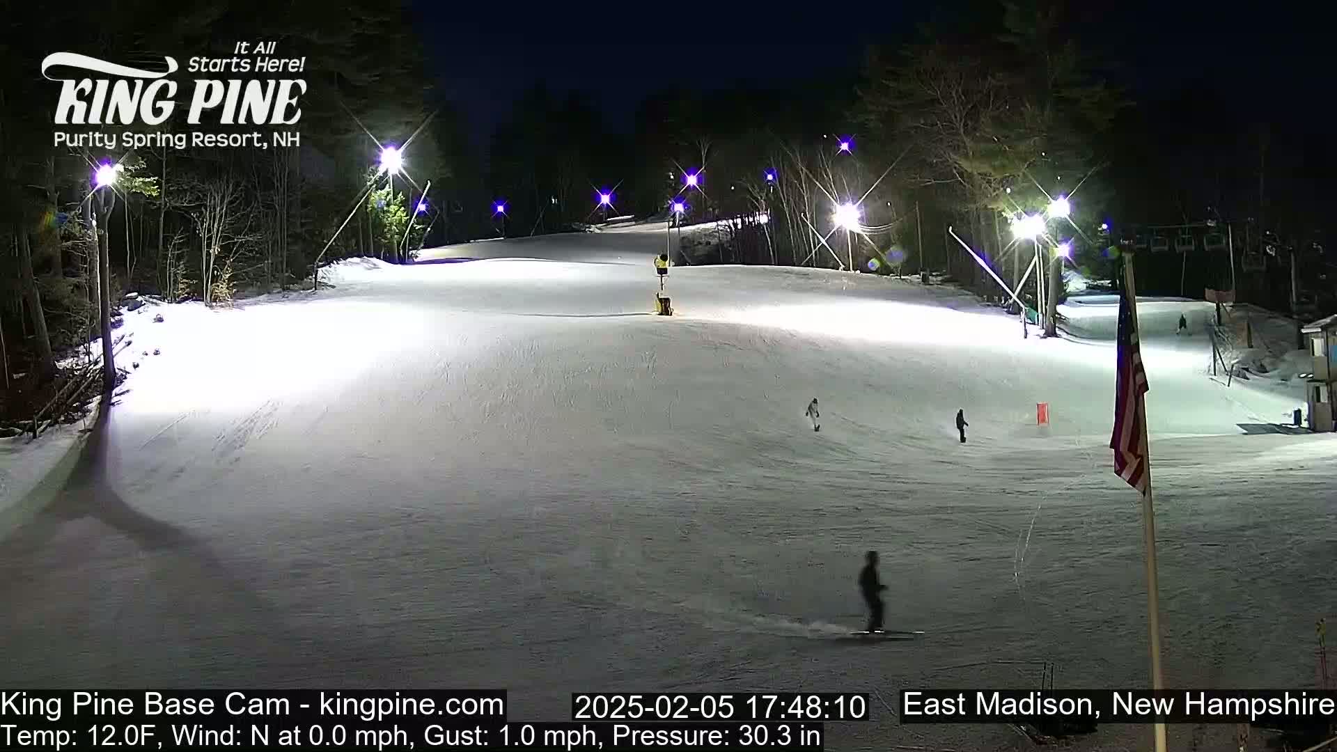 A snow-covered ski slope is illuminated at night by several lights, with a few skiers visible on the slope.