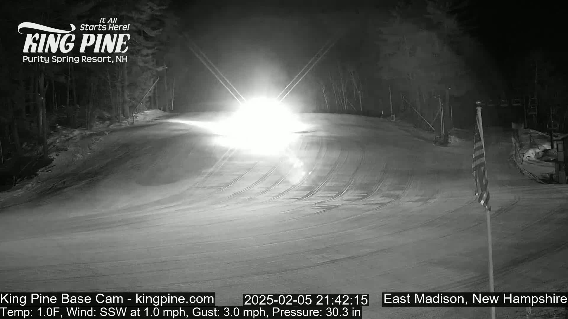 A snow-covered ski slope at night is illuminated by bright headlights from a vehicle approaching from the center of the frame, under a dark, clear sky.