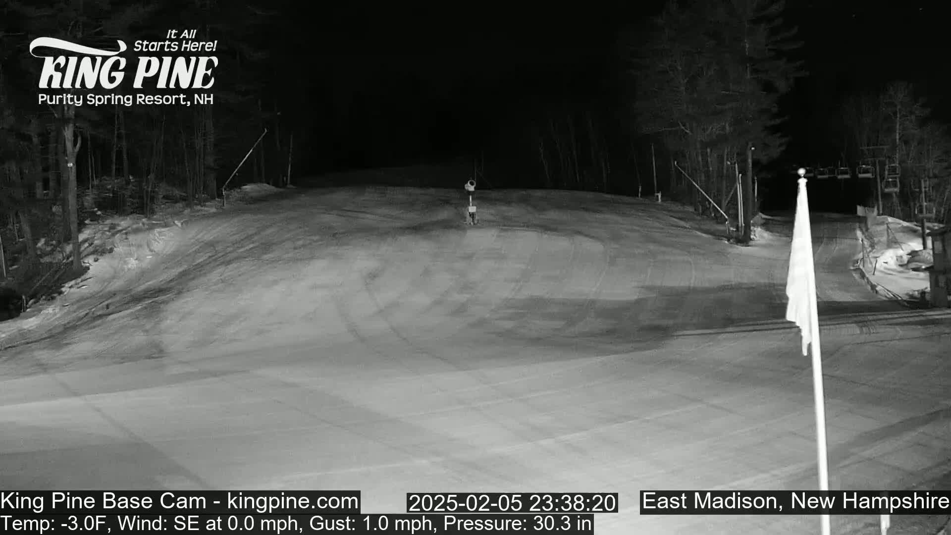 A snow-covered ski slope at night with a few trees visible in the background under a clear, dark sky.