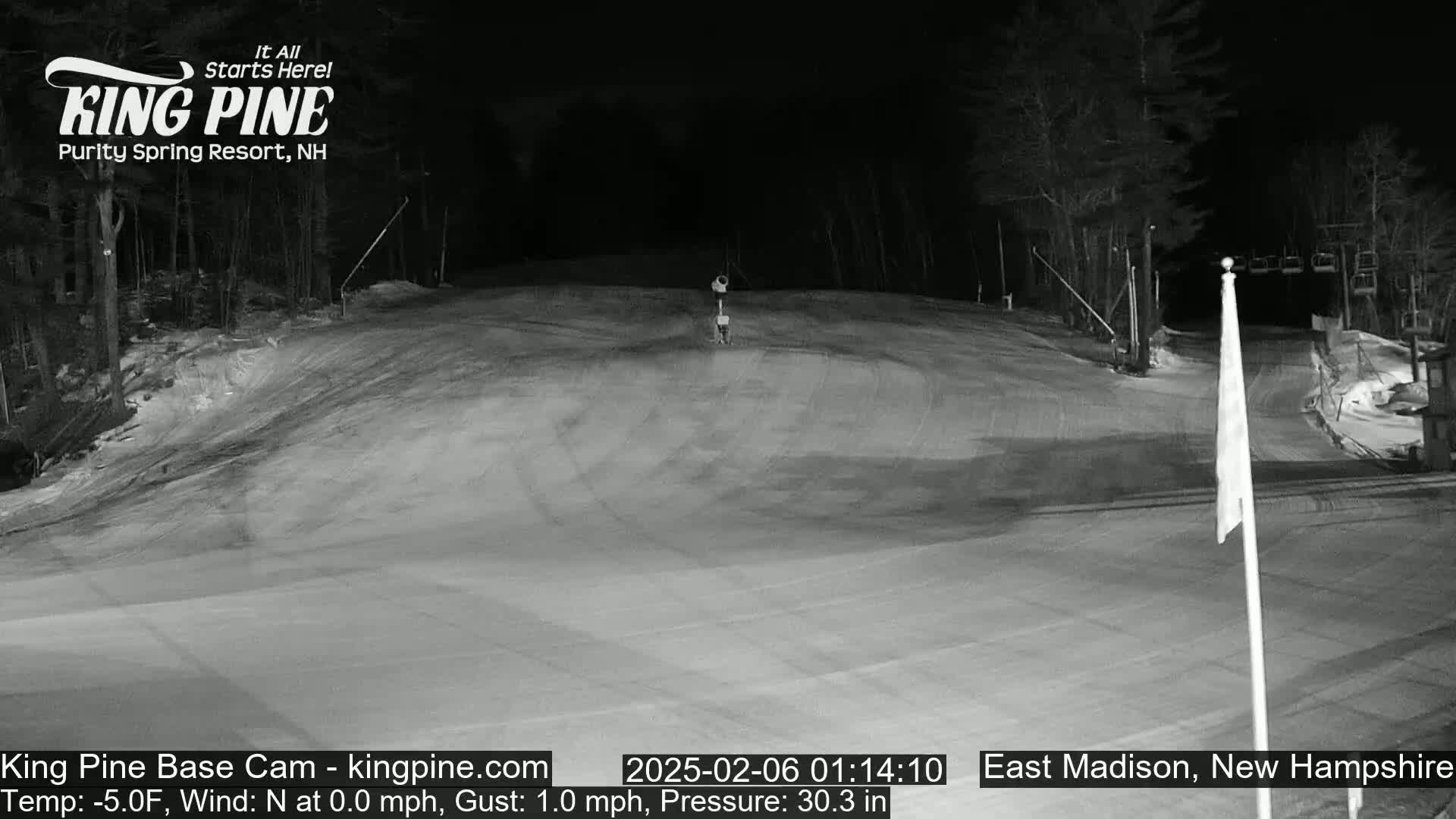A snow-covered ski slope at night, illuminated by artificial light, with trees in the background and a calm wind.