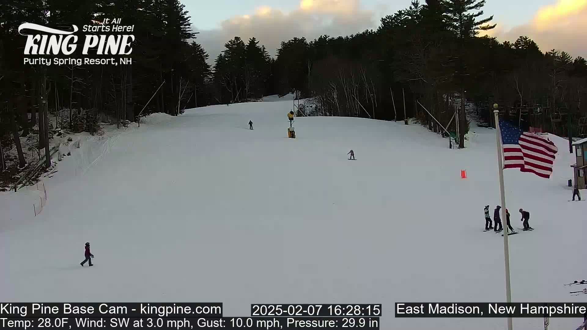 A snow-covered ski slope with several skiers and snowboarders, under a partly cloudy sunset sky.