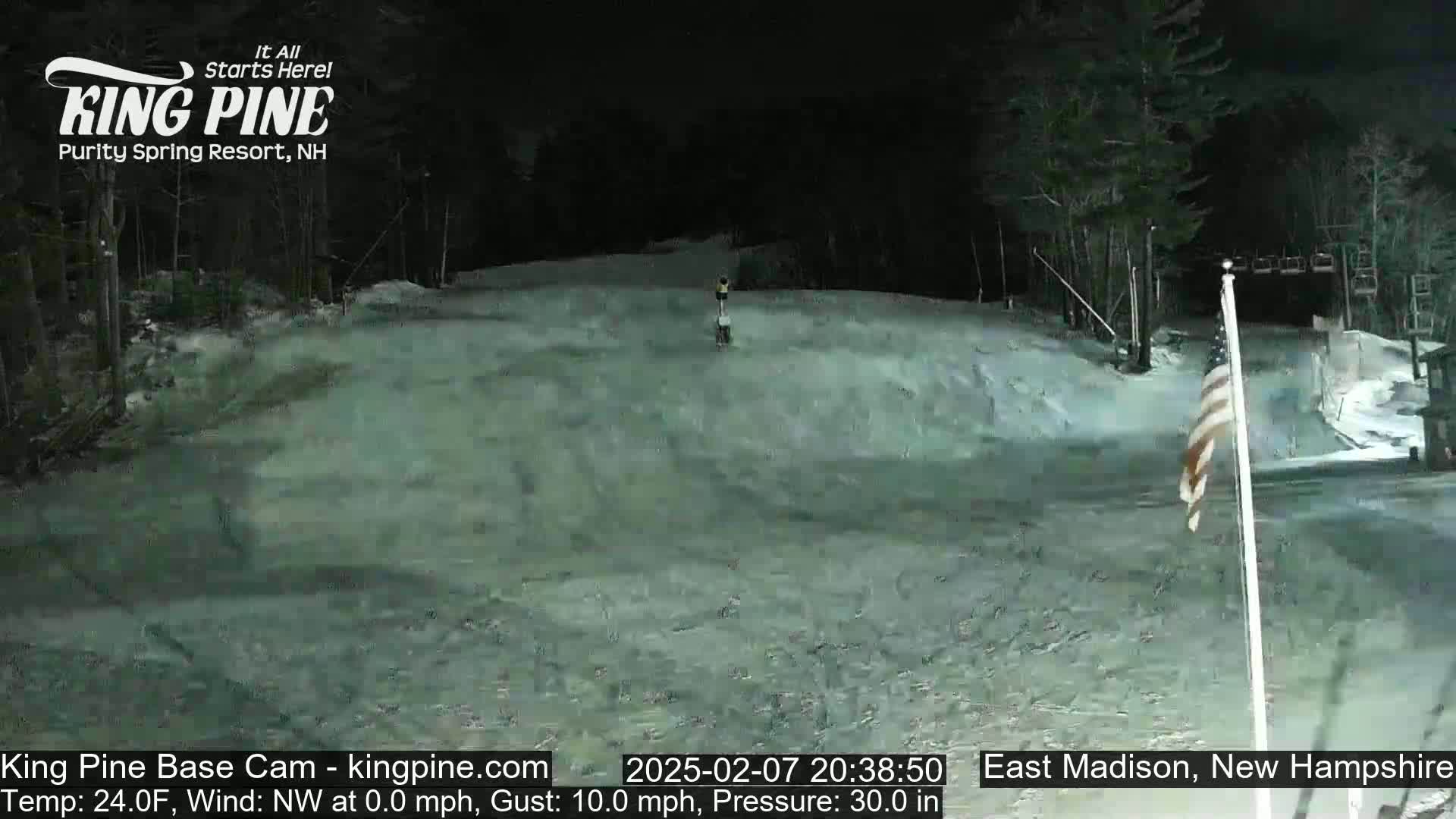 A nighttime view of a snow-covered ski slope with a single person skiing down, under a dark sky.