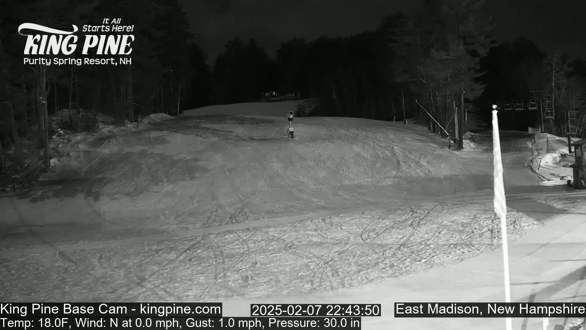 A snow-covered ski slope at night, illuminated by artificial light, with trees in the background under a dark sky.