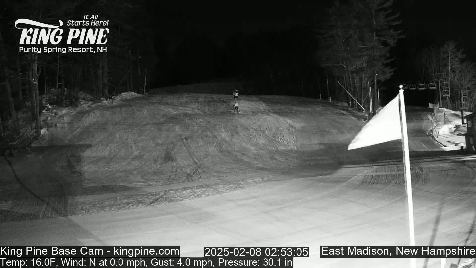 A snow-covered ski slope at night, illuminated by artificial light, with calm winds.