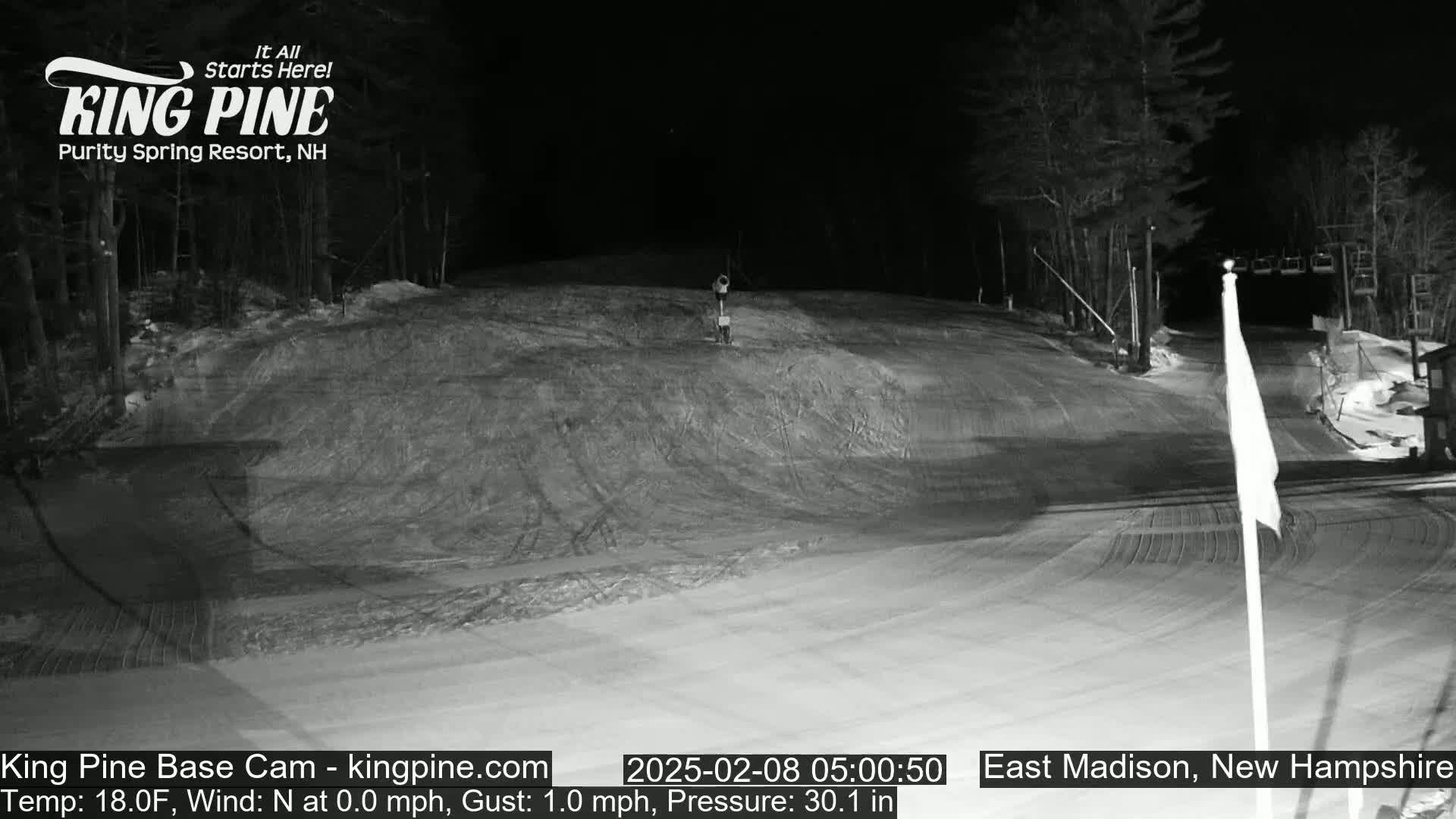 A snow-covered ski slope at night, illuminated by artificial light, with trees in the background and a flagpole in the foreground.
