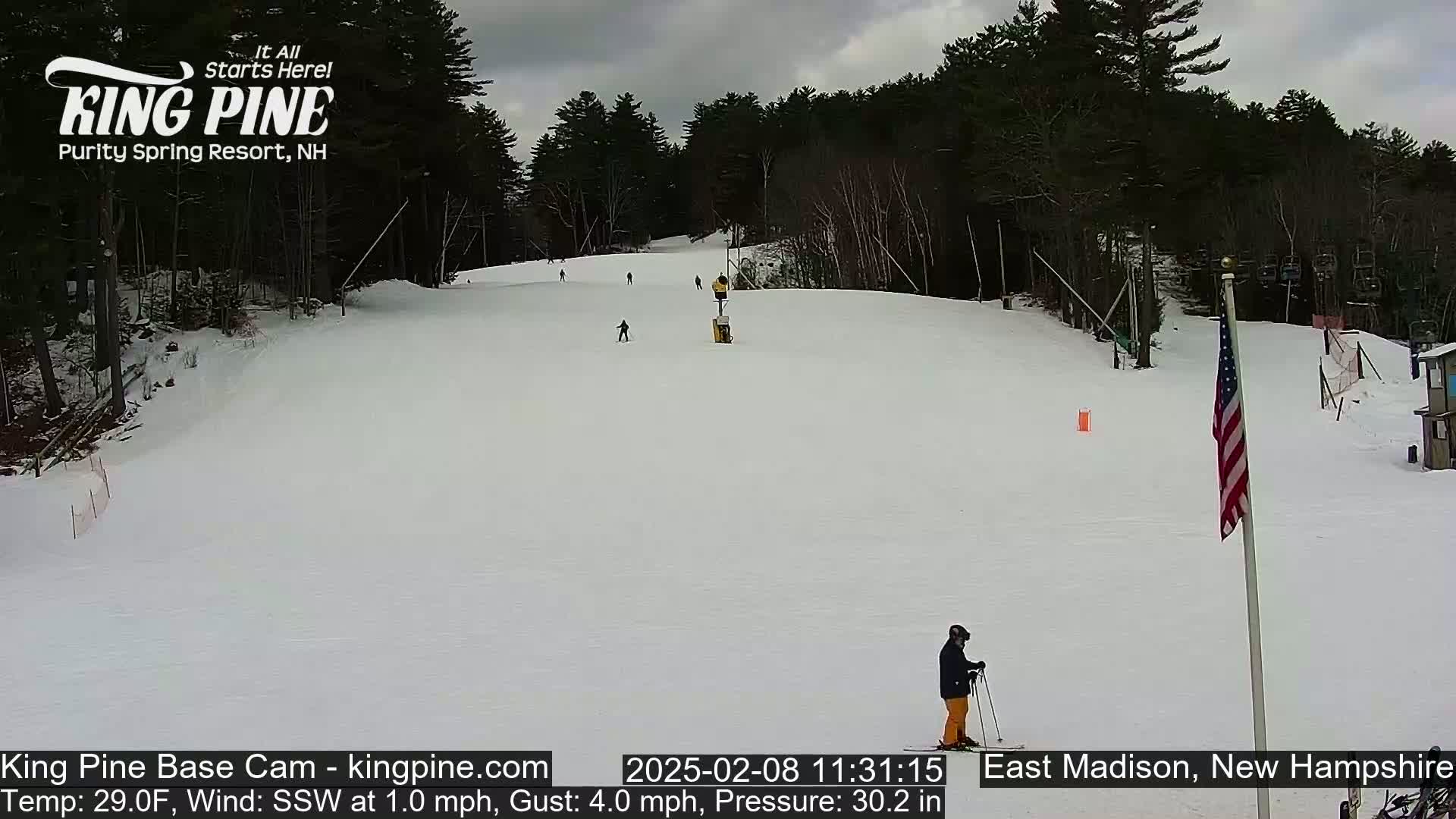 A snowy ski slope with several skiers, a lift, and an American flag under overcast skies.