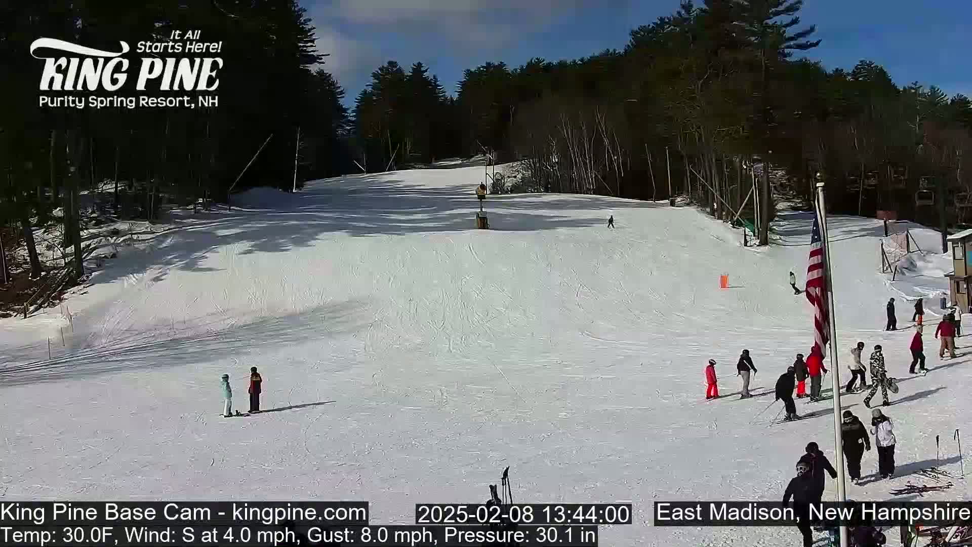 A sunny day at a snow-covered ski resort shows skiers and snowboarders on a slope surrounded by trees.