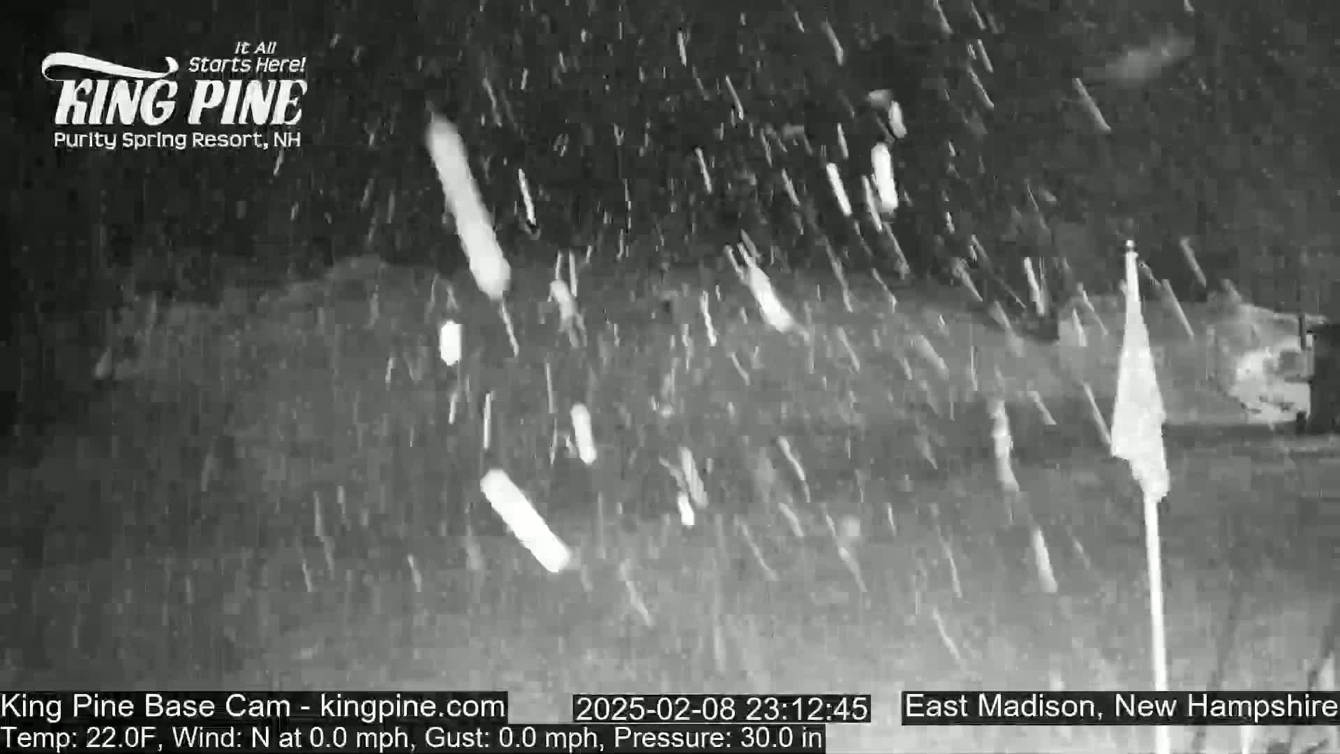 A nighttime, snowy scene shows falling snow and a flagpole with a blank flag.