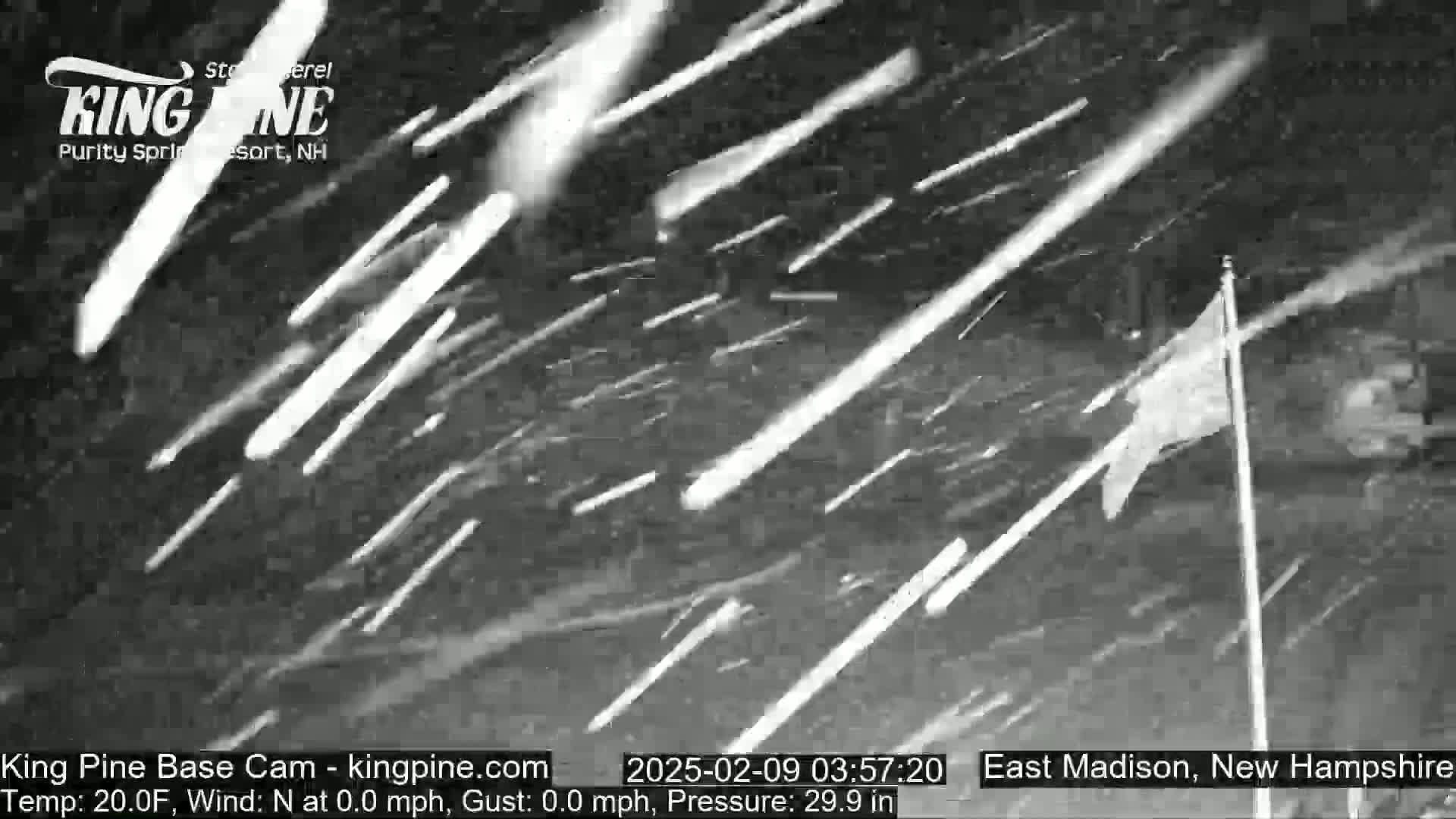 A nighttime outdoor scene shows heavy snowfall with streaks of bright light from falling snow illuminated by a light source, and a flagpole with a partially visible flag in the lower right.