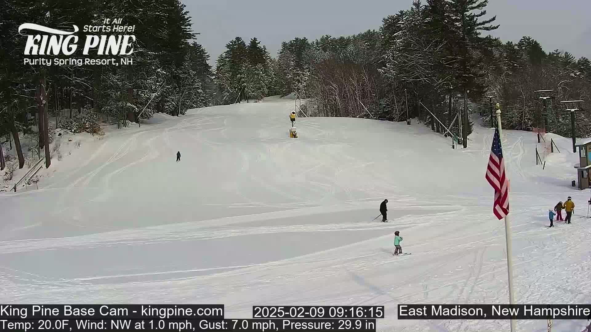 A snow-covered ski slope with several skiers and a snowmaking machine, under a cloudy sky.