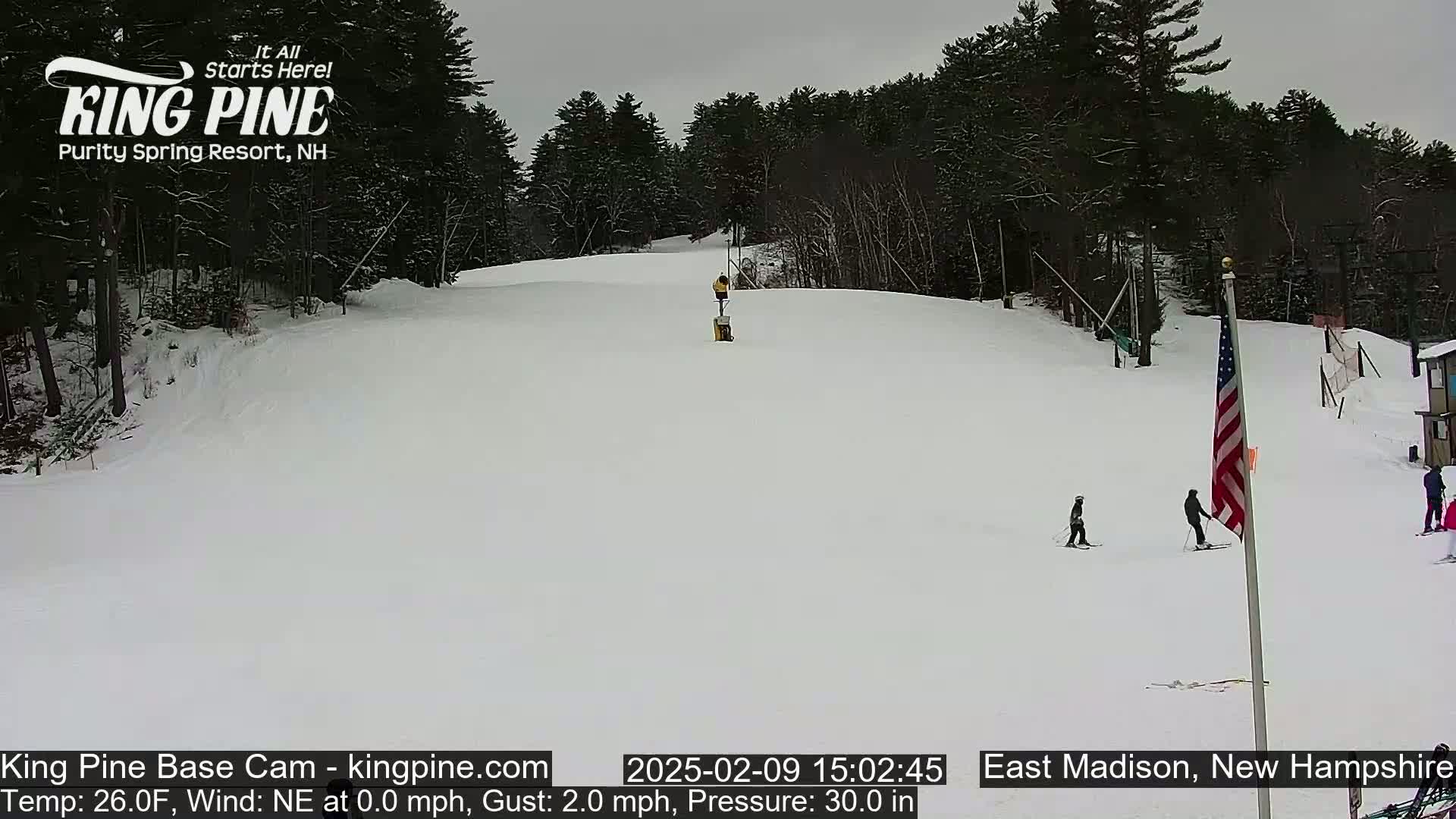 A snow-covered ski slope with several skiers visible, under an overcast sky.