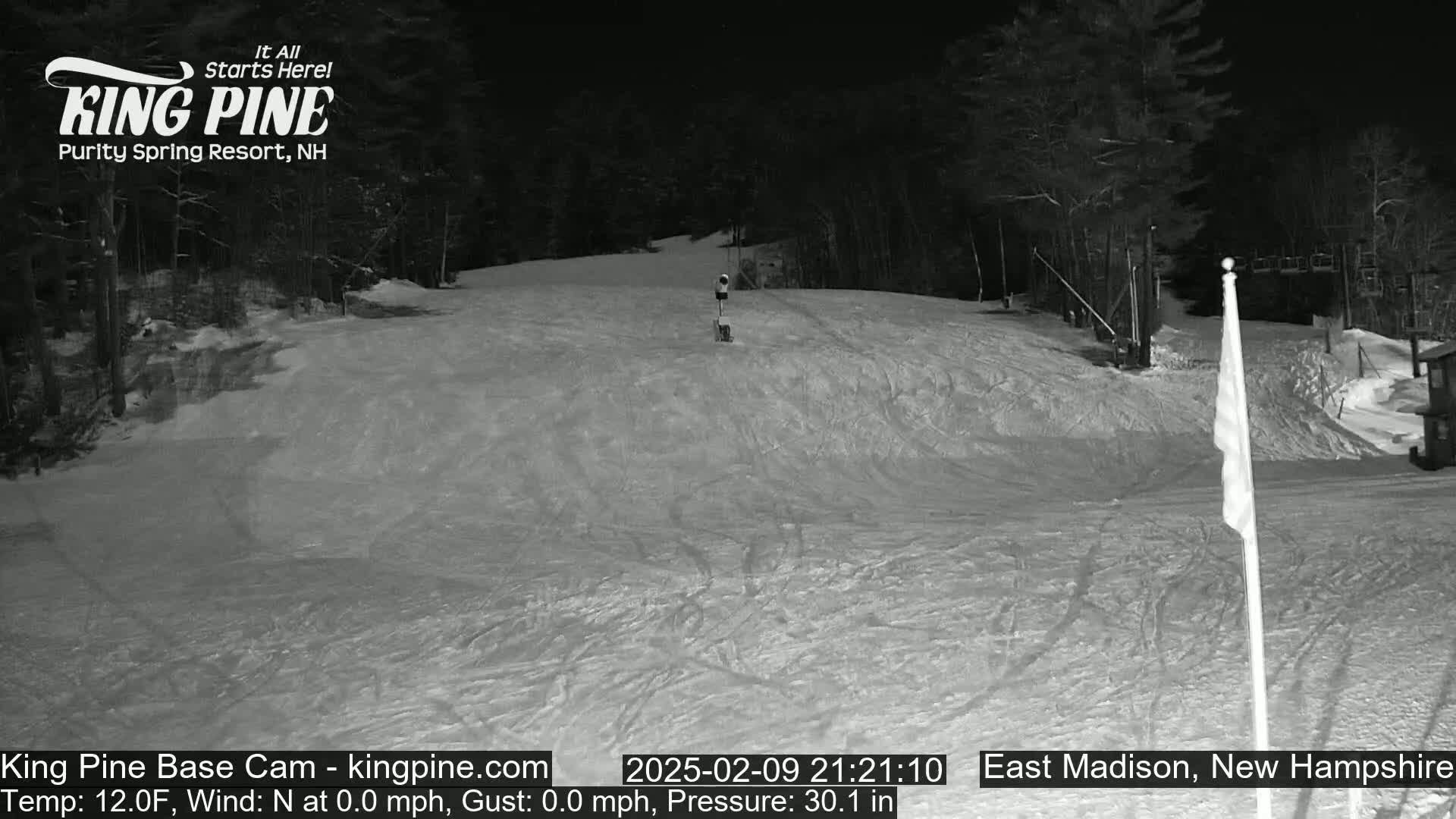 A snow-covered ski slope at night, illuminated by artificial light, with a person visible in the distance and trees lining the background.