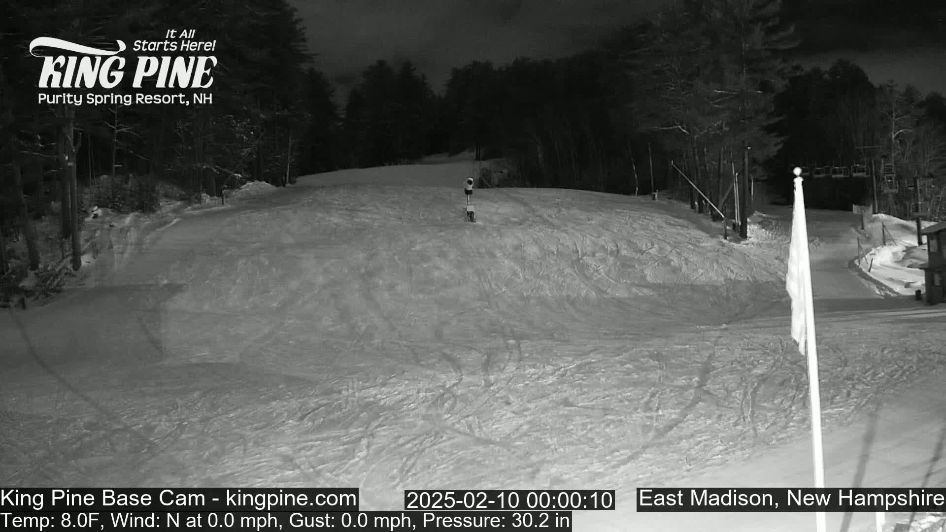 A snow-covered ski slope at night, illuminated by artificial light, with trees in the background and a calm, cold temperature.