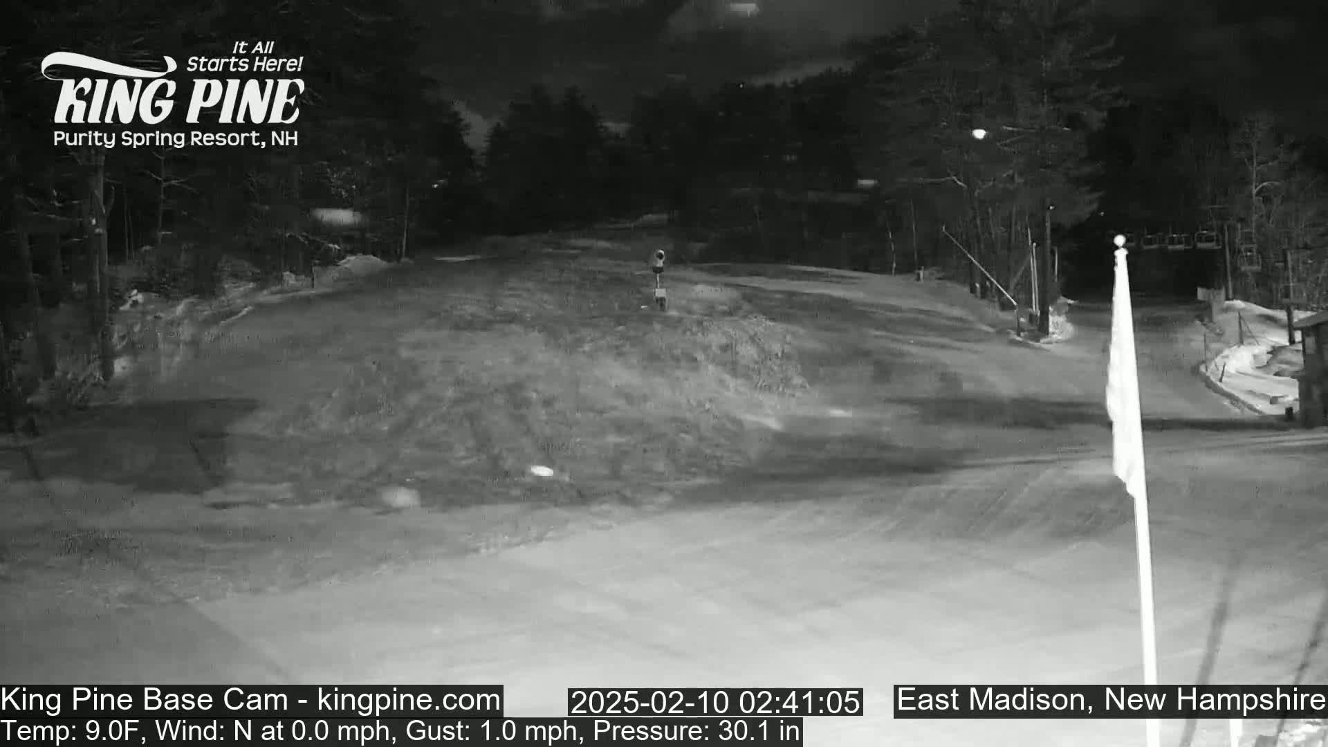 A nighttime view of a snow-covered area, possibly a ski resort, with trees in the background under a dark sky.