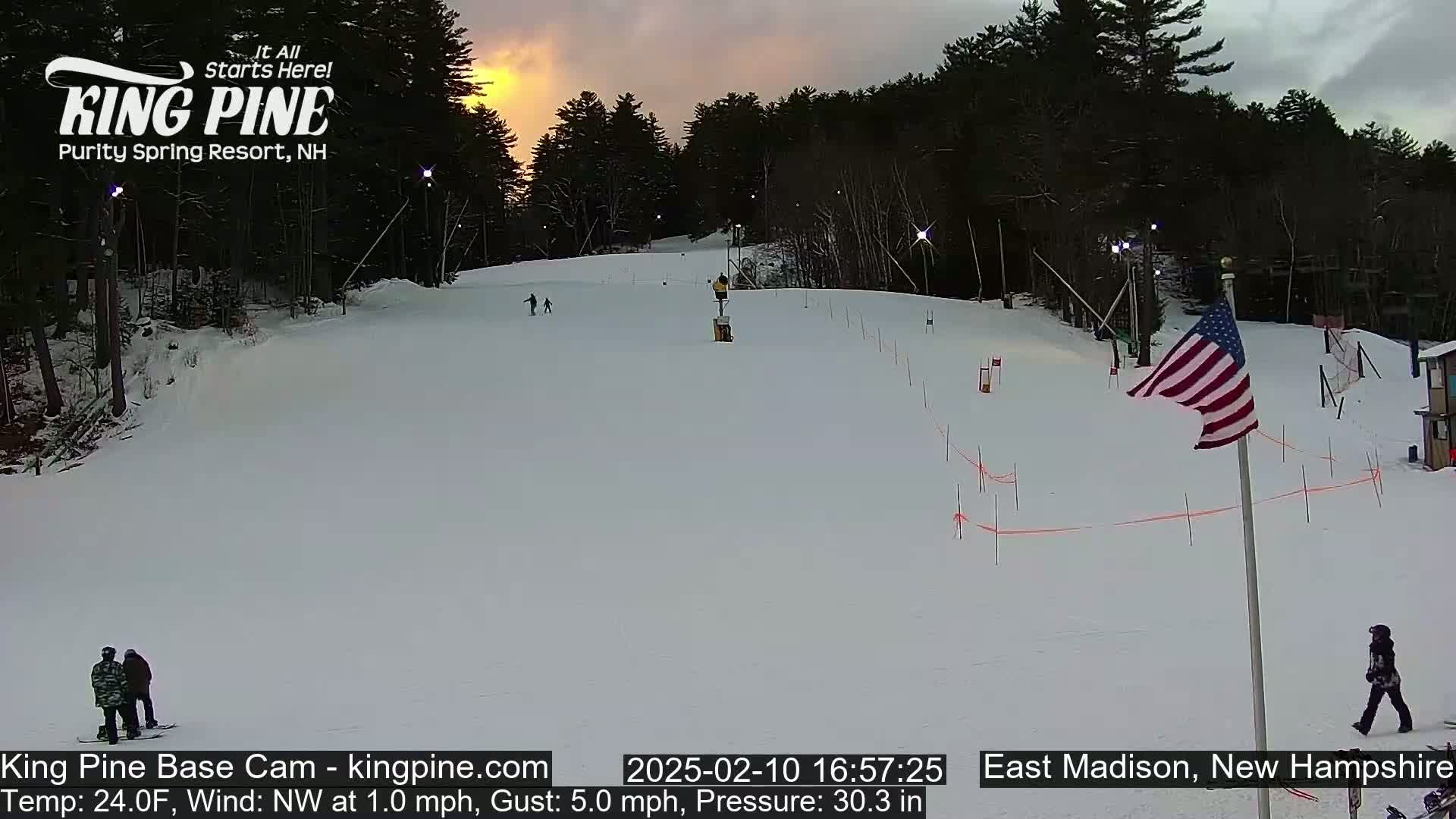 A snow-covered ski slope at sunset with several skiers and snowboarders, a small American flag, and a few people walking near a building.