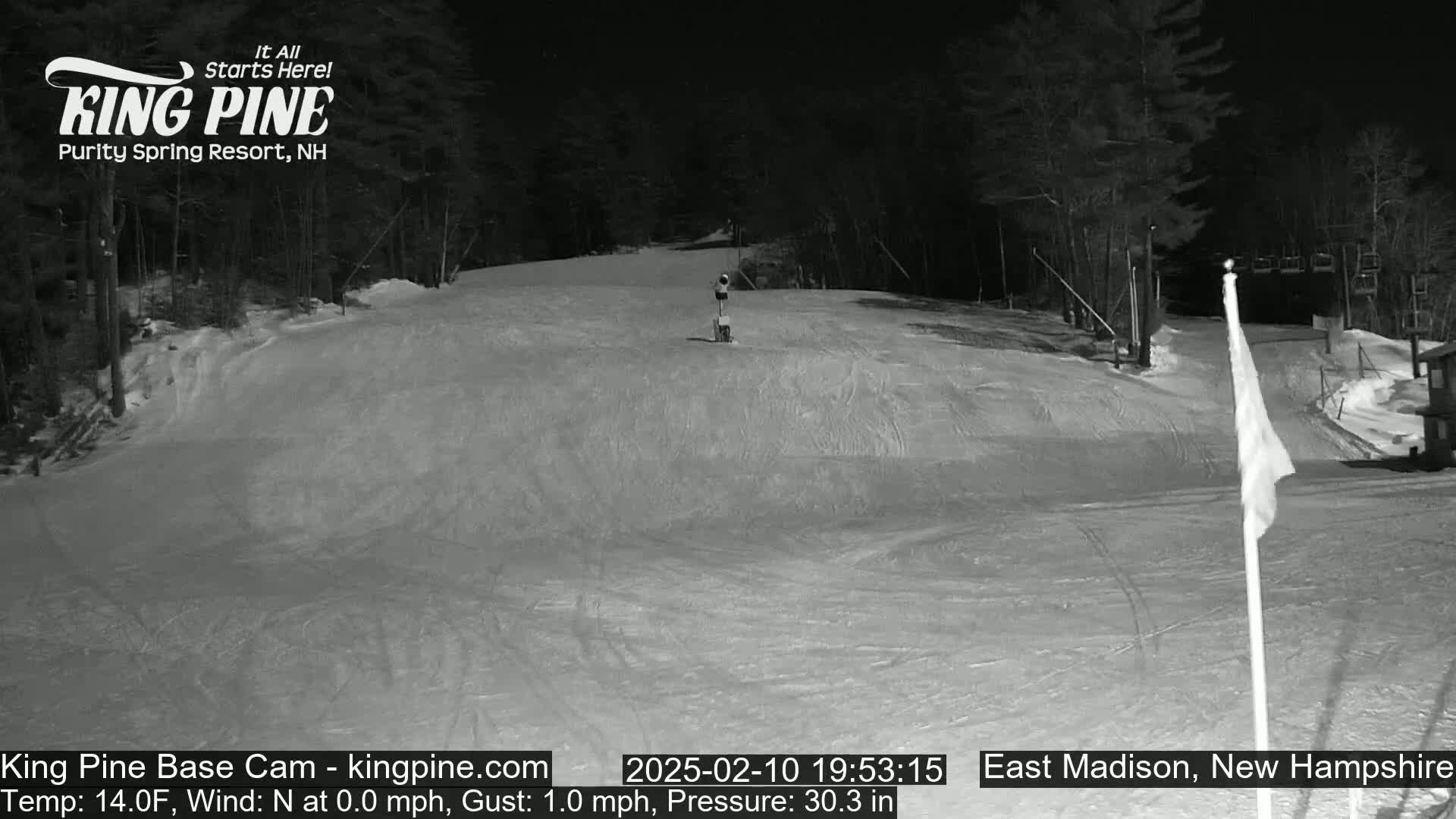 A snow-covered ski slope at night is illuminated, with a calm wind and trees lining the edges.