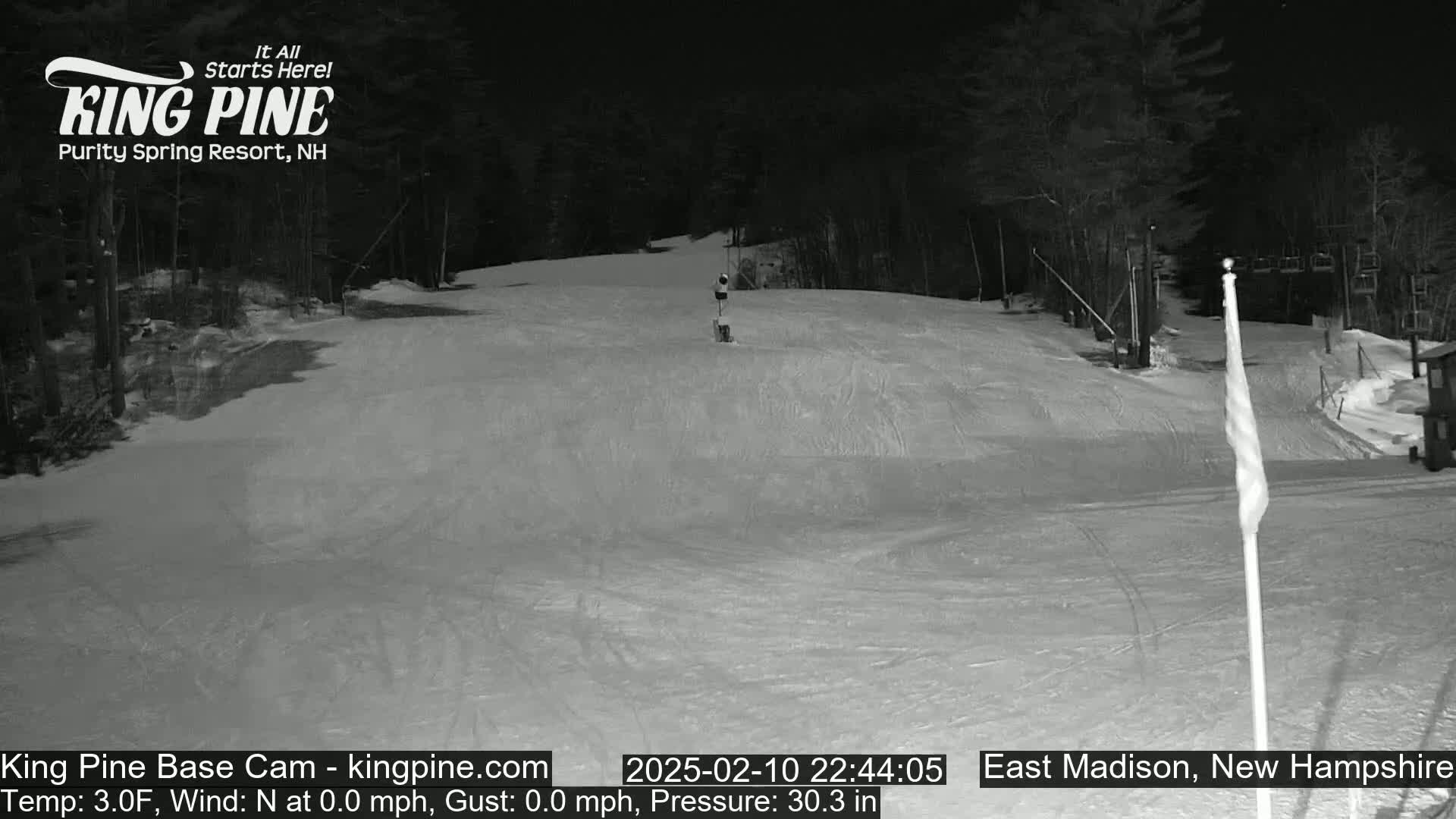 A nighttime, snow-covered ski slope is illuminated, showing a mostly empty run with a few tracks and a ski lift in the background.