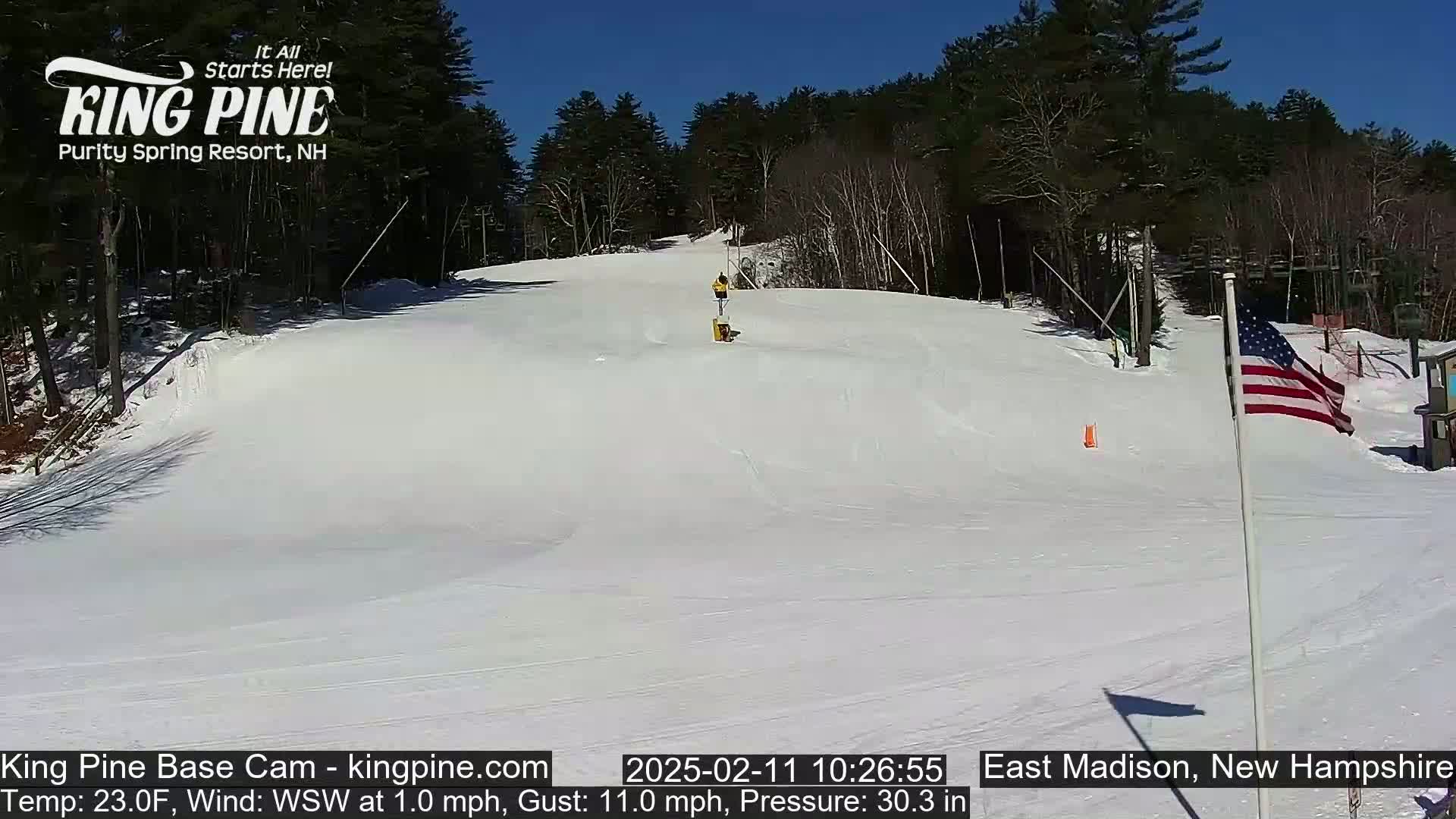 A snow-covered ski slope is visible under a bright, sunny sky, with a person in the distance on the slope, and an American flag standing nearby.