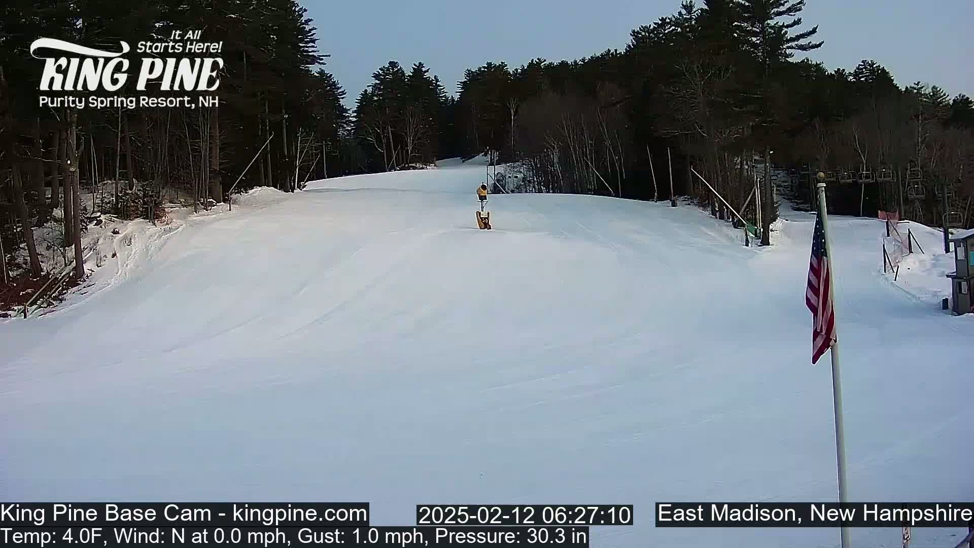 A snow-covered ski slope with a person on it, under a clear sky, is surrounded by trees.