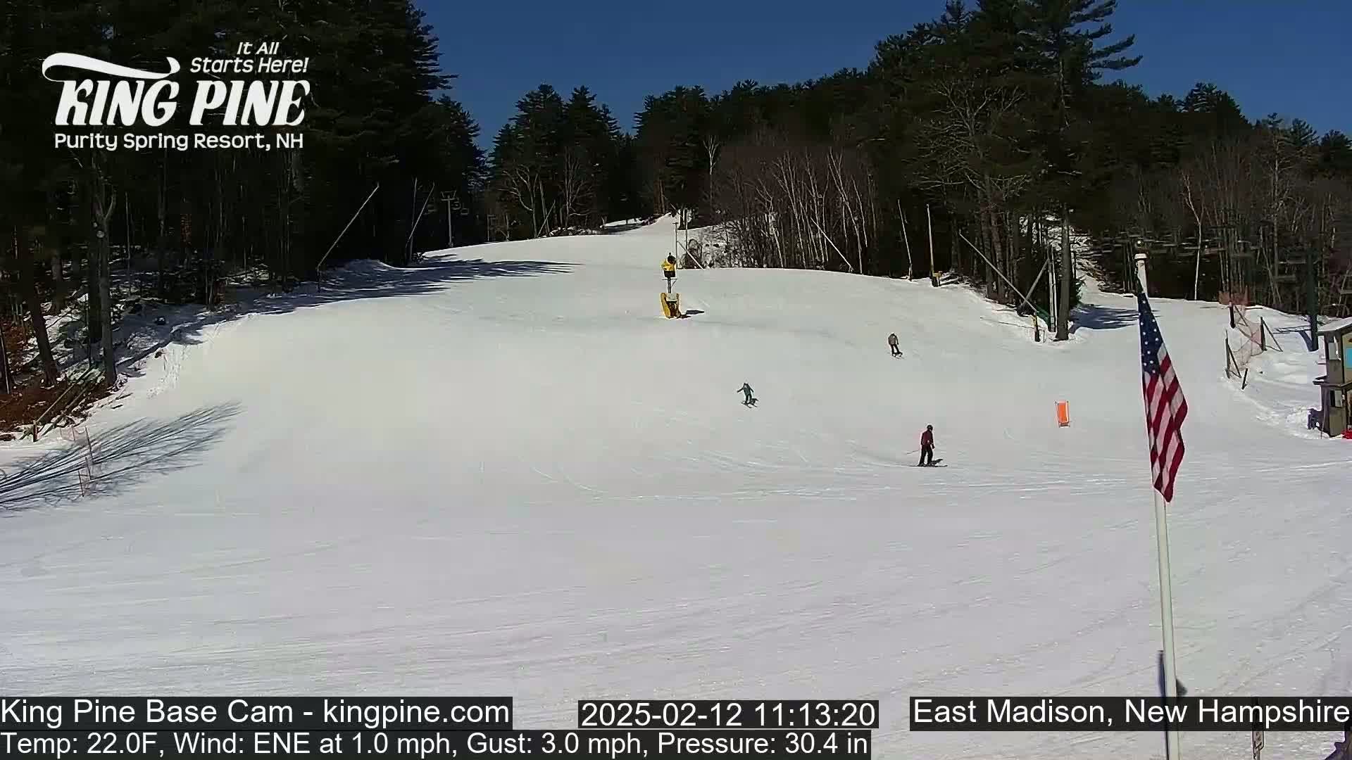 A snow-covered ski slope with several skiers, a ski lift, and a US flag on a sunny day.
