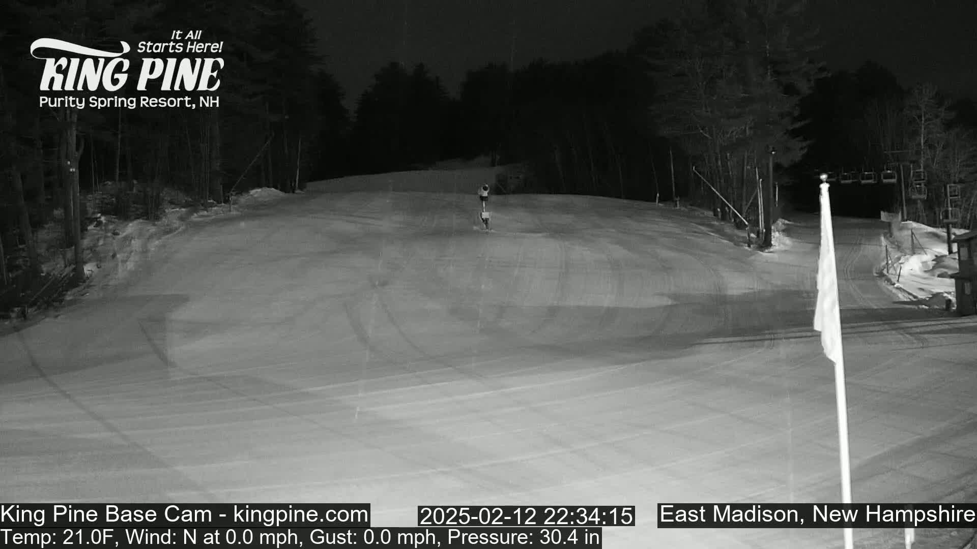 A mostly empty snow-covered ski slope at night, illuminated by artificial light, with a ski lift visible in the background.
