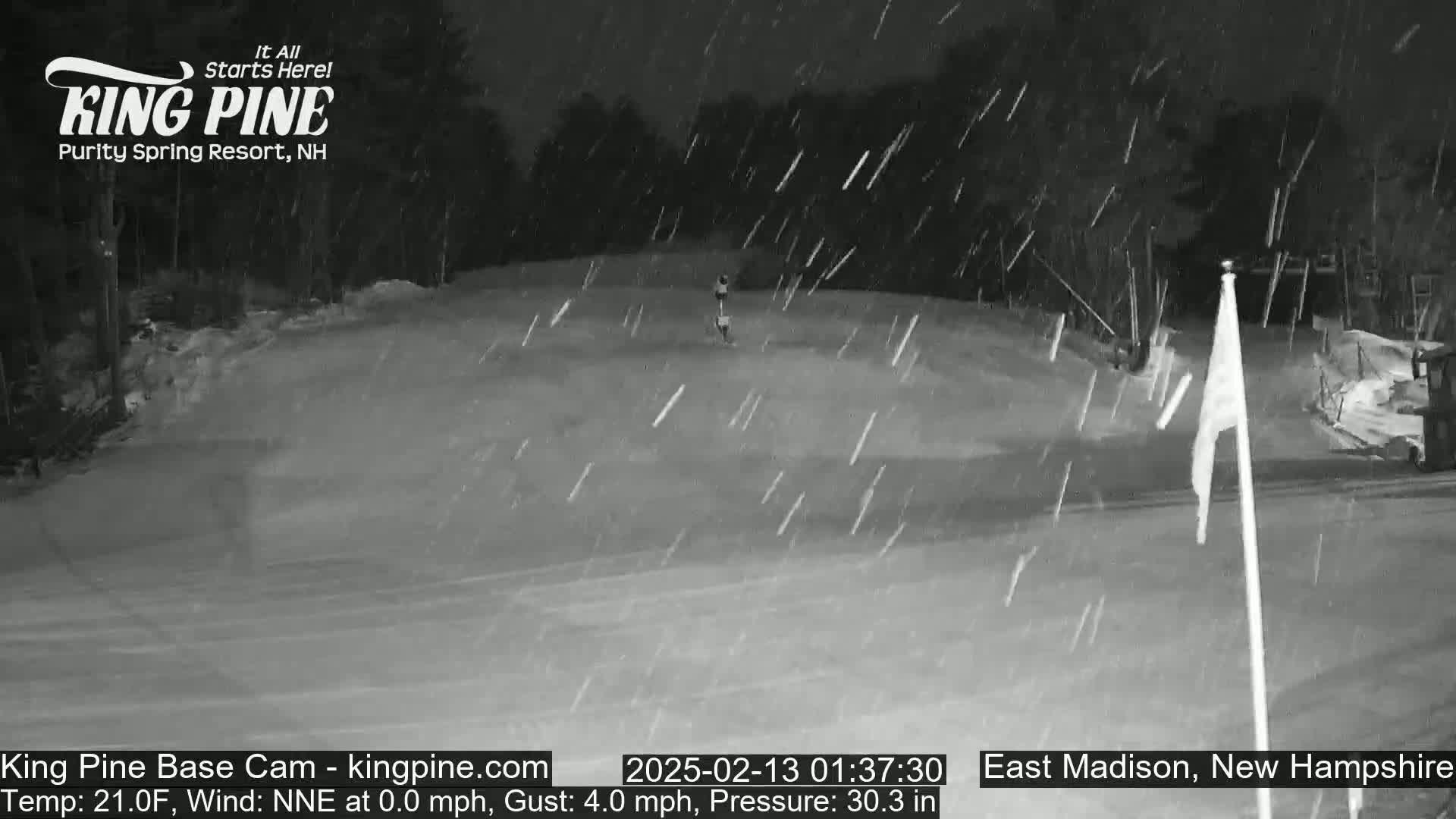 A snowy nighttime scene shows a mostly empty snow-covered area with a flagpole and trees in the background under falling snow.