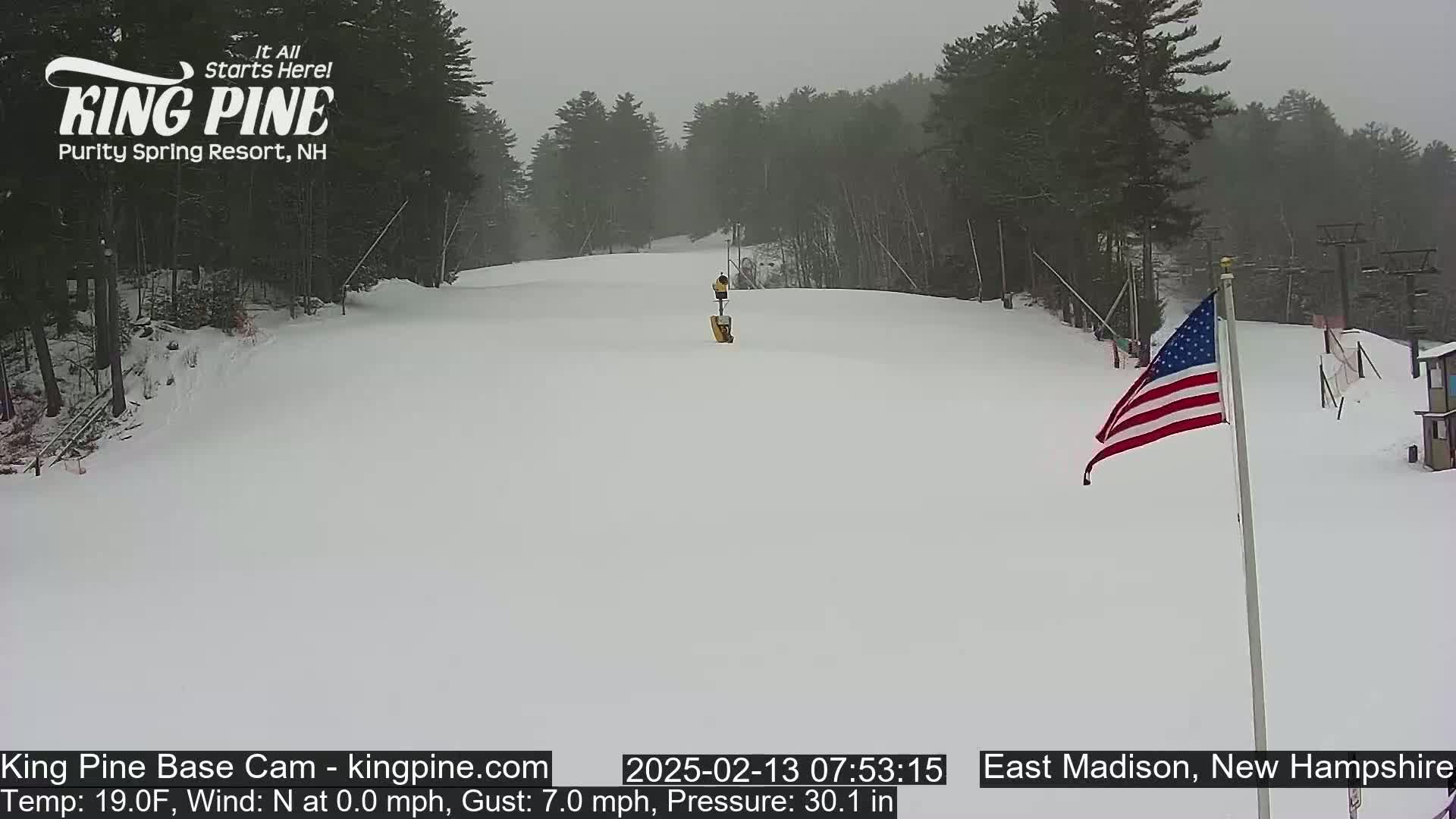 A snow-covered ski slope is visible on an overcast day, with a lone figure near a lift and an American flag in the foreground.