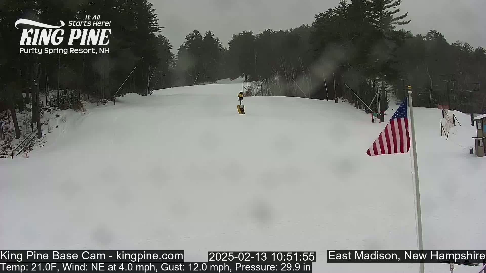 A snowy ski slope with a person skiing down it, under an overcast sky.