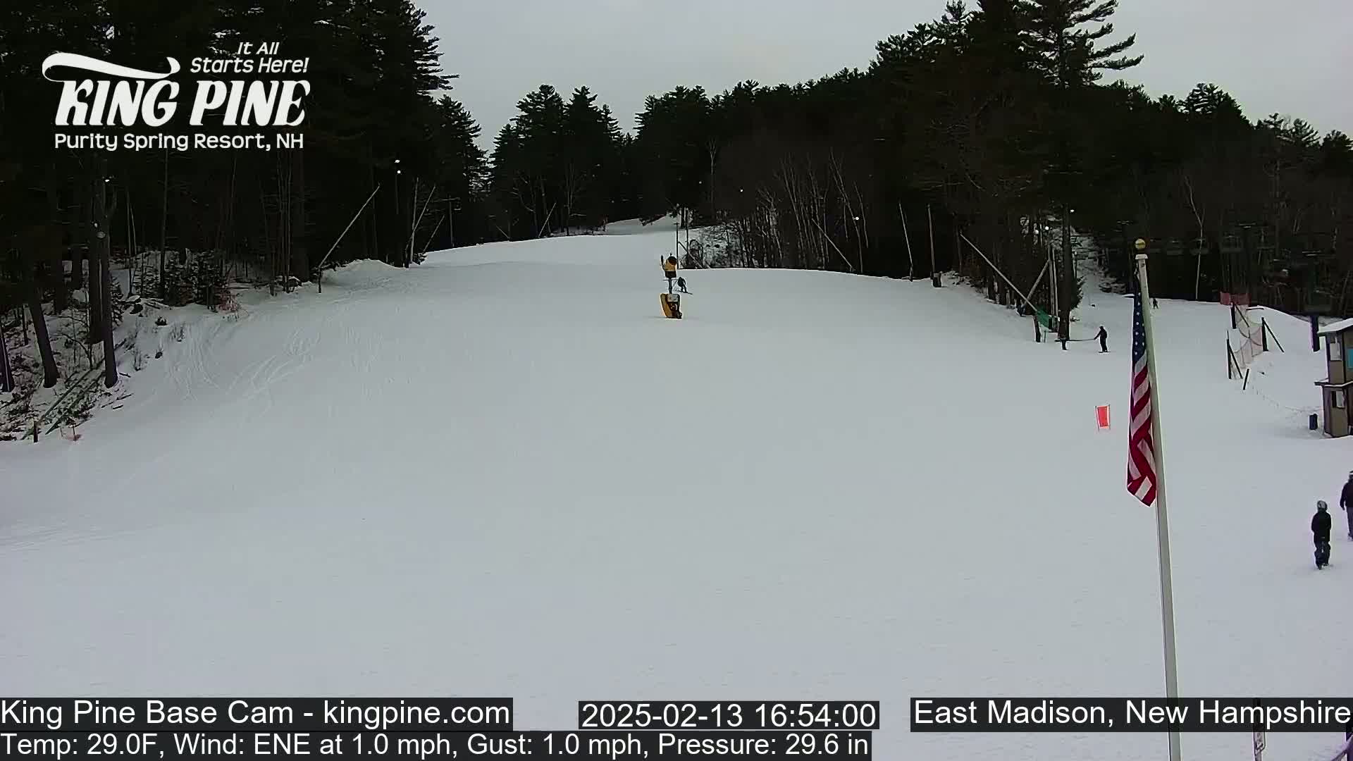 A snowy ski slope with several skiers and a chairlift, under an overcast sky.