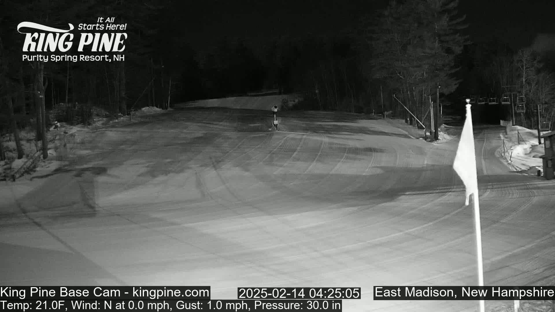 A snow-covered ski slope at night, illuminated by artificial light, with a calm wind.