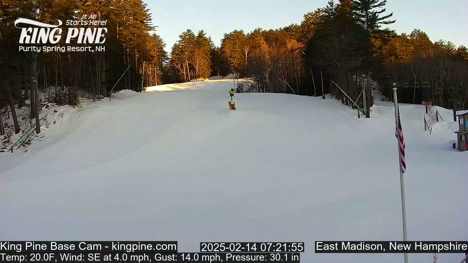 A snow-covered ski slope with a person skiing down it, under a clear, sunrise sky, with trees lining the slope and a small American flag in the foreground.