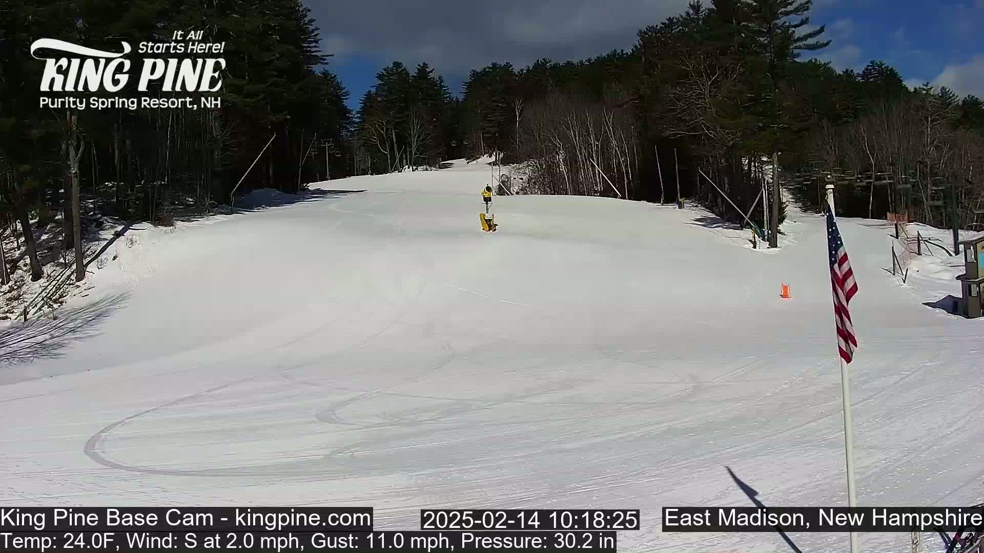 A snowy ski slope with a person on it is visible under a sunny sky, surrounded by trees.